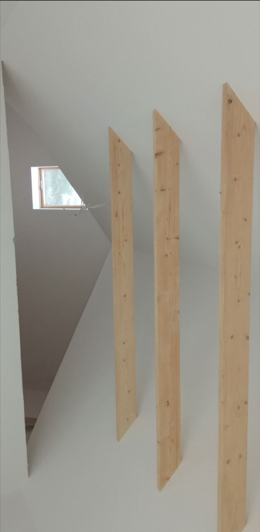 Skośny sufit z oknem i drewnianymi elementami dekoracyjnymi. Trzy pionowe, drewniane belki o ściętych końcach przymocowane do ściany. Minimalistyczny design.