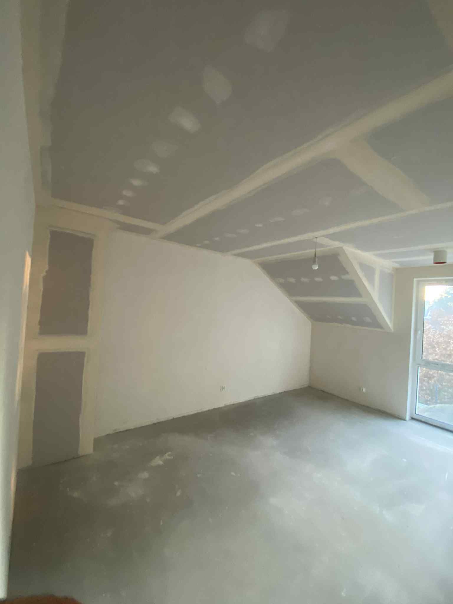 Pokój ze skośnym sufitem po montażu płyt gipsowo-kartonowych, widoczne łączenia i szpachlowane miejsca. Surowy beton na podłodze, okno z widokiem na zewnątrz.