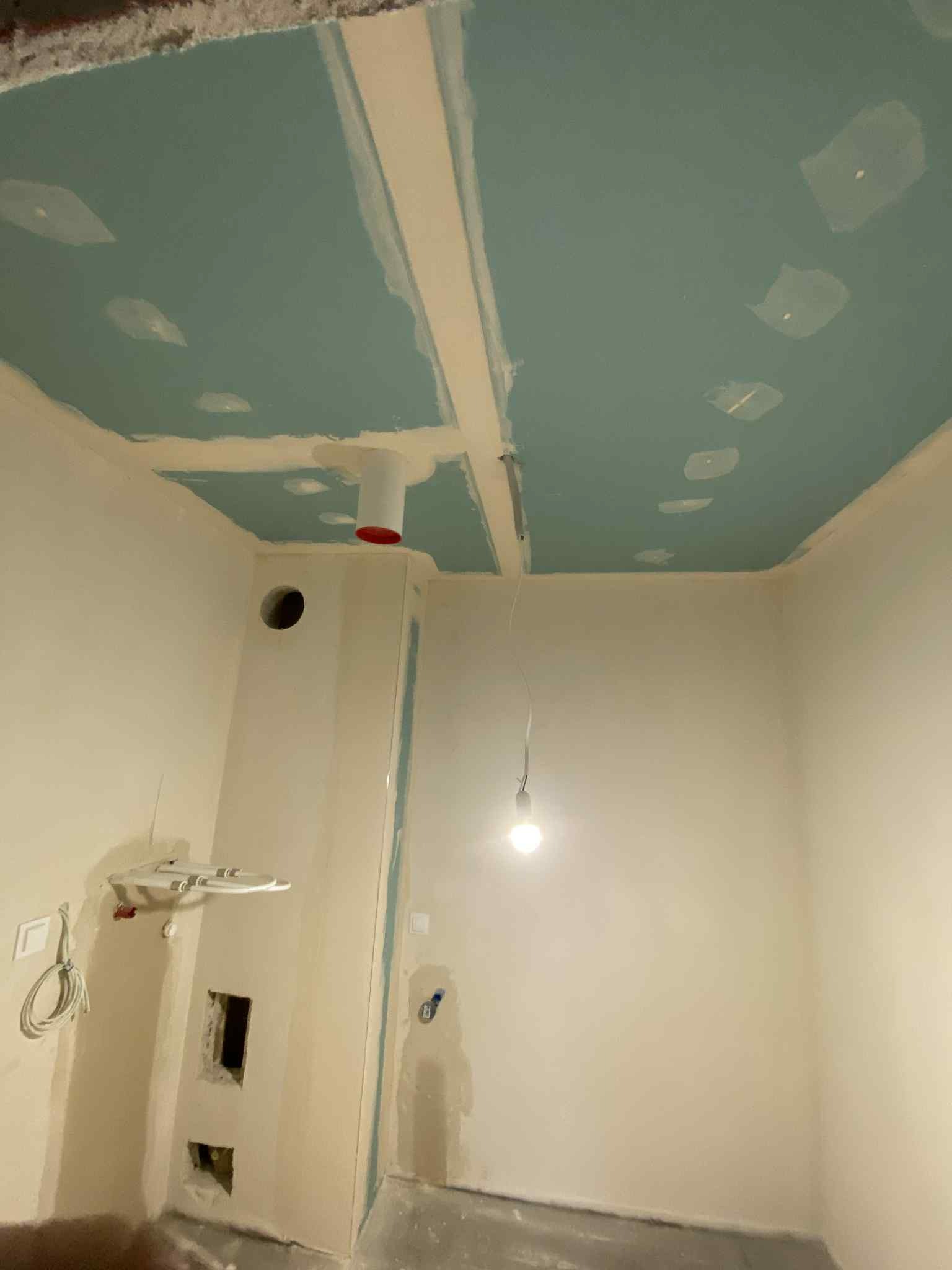 Narożne wnętrze w trakcie prac wykończeniowych: sufit z płyt kartonowo-gipsowych, częściowo wyszpachlowany, ściany przygotowane pod malowanie, widoczne instalacje.