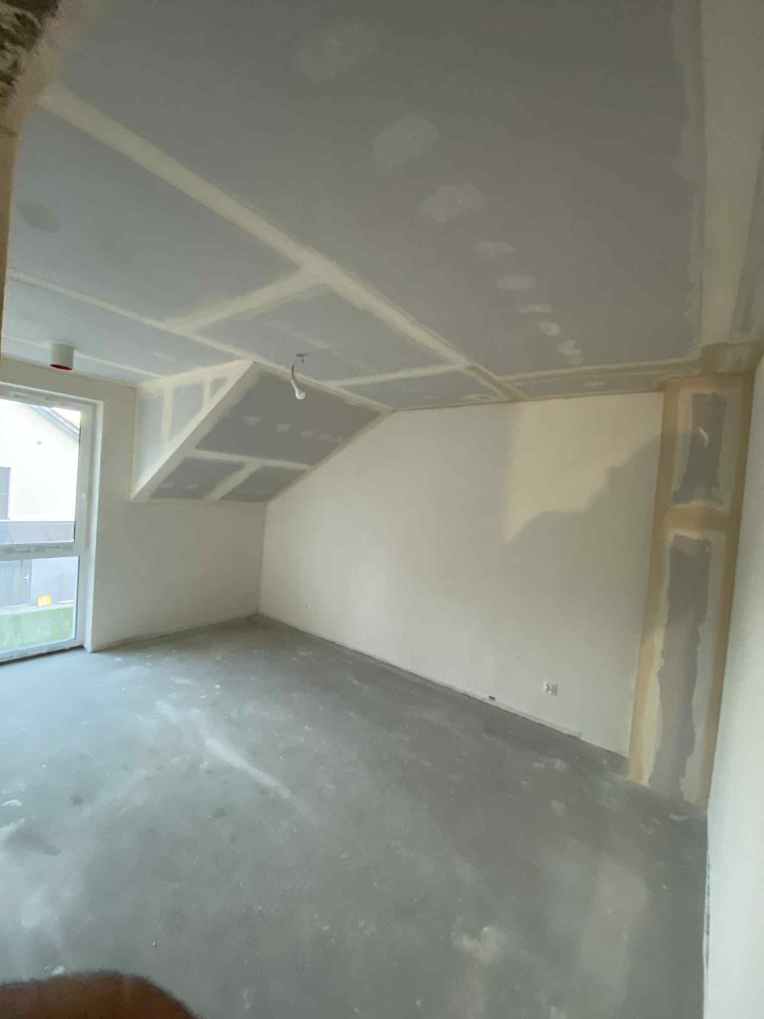 Surowe wnętrze z płytami gipsowo-kartonowymi na suficie i ścianach, przygotowane do dalszych prac wykończeniowych. Widoczne łączenia płyt i surowa podłoga.