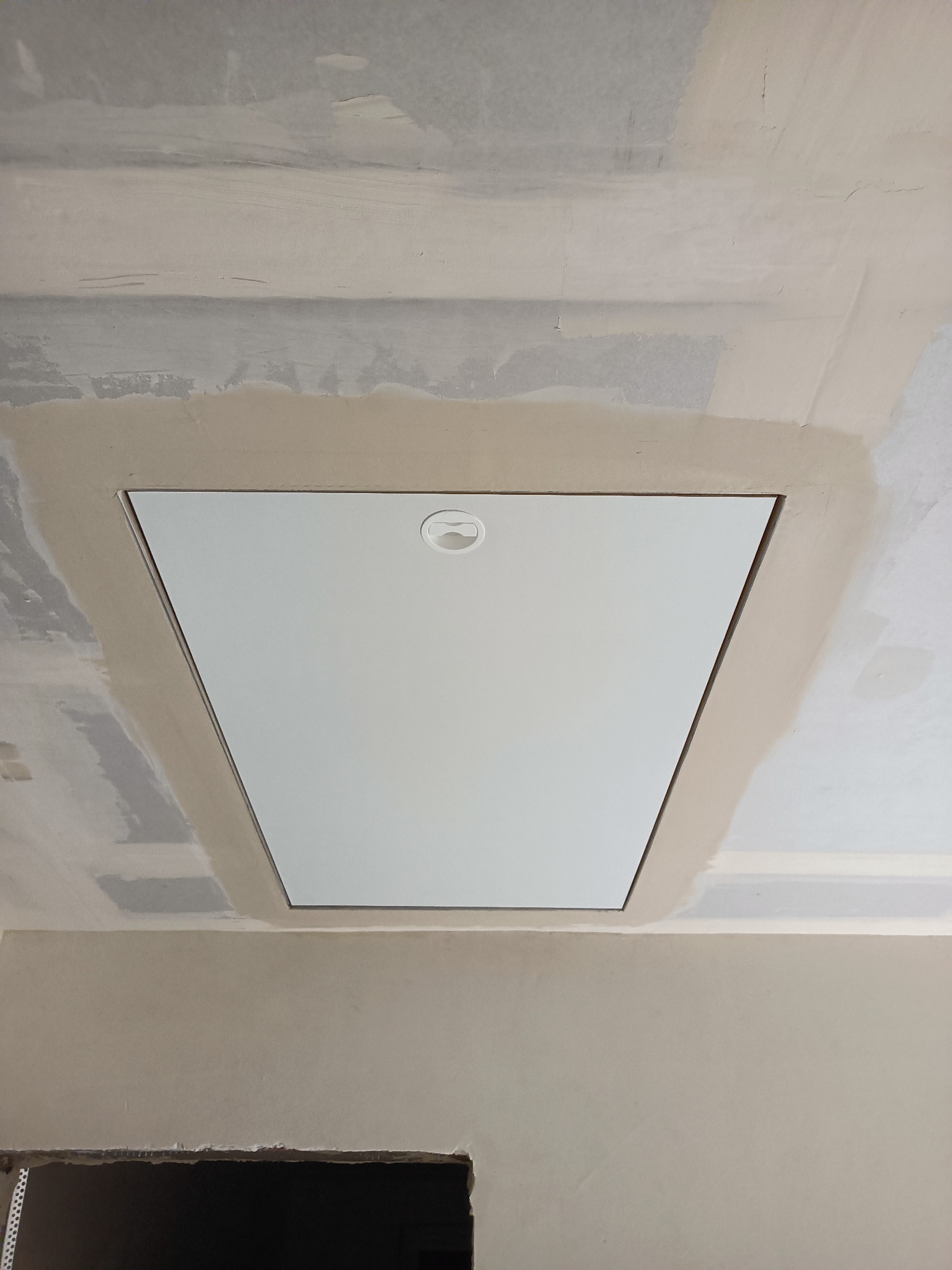 Biała klapa rewizyjna w suficie, otoczona świeżo nałożoną warstwą szpachli. Widoczne niedoskonałości i surowość wykończenia sufitu. Fragment ściany w dolnej części kadru.