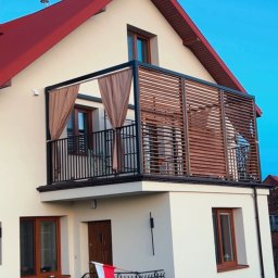 GOR-WELD Krzysztof Gorlewski - Dom z balkonem z metalową balustradą i drewnianą pergolą z zasłonami. Na schodach flaga Polski. Ujęcie z dołu, błękitne niebo w tle.
