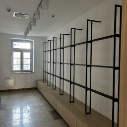 GOR-WELD Krzysztof Gorlewski - Minimalistyczne, czarne, metalowe regały na jasnym tle ściany, zamontowane nad drewnianą zabudową. Widok na wnętrze z oknem i grzejnikiem.