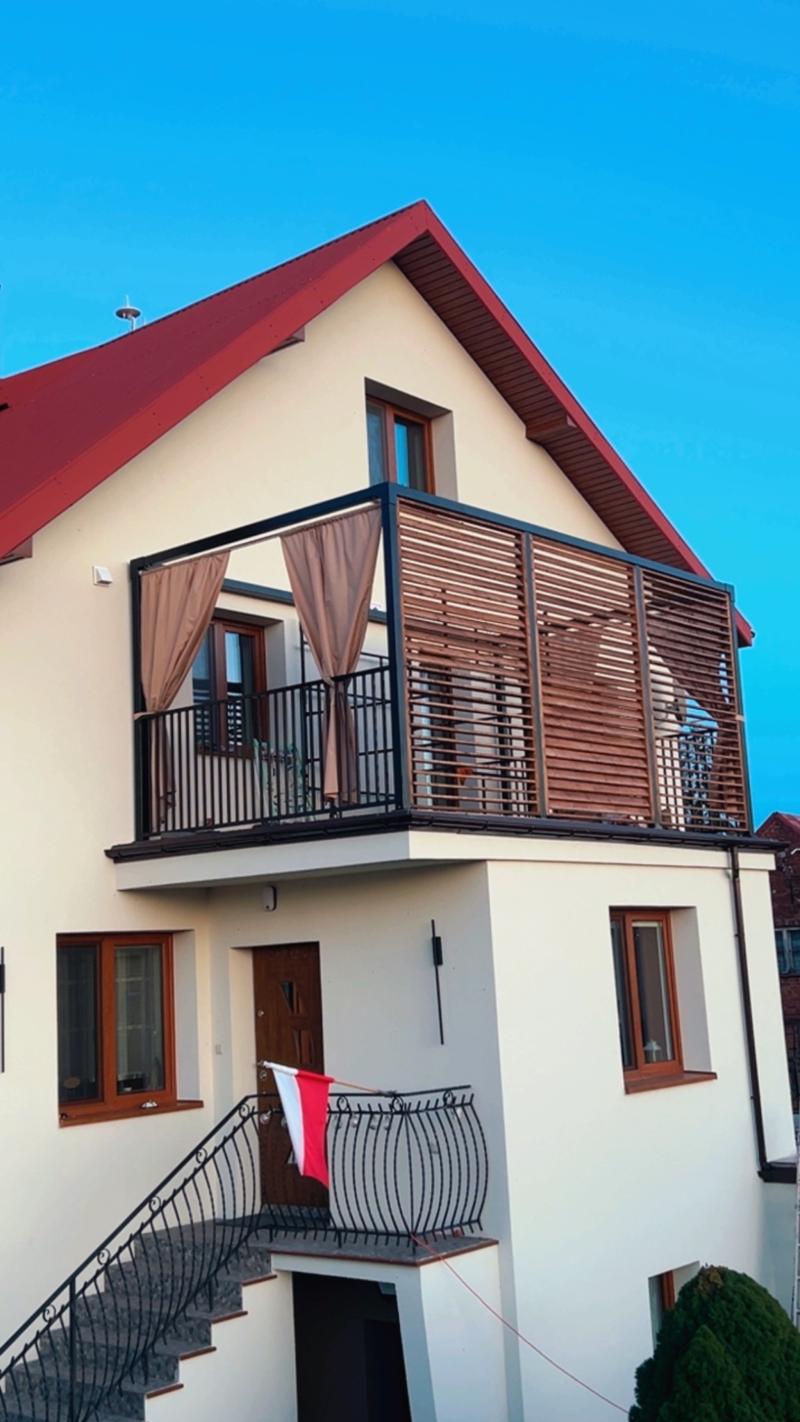 Dom z balkonem z metalową balustradą i drewnianą pergolą z zasłonami. Na schodach flaga Polski. Ujęcie z dołu, błękitne niebo w tle.