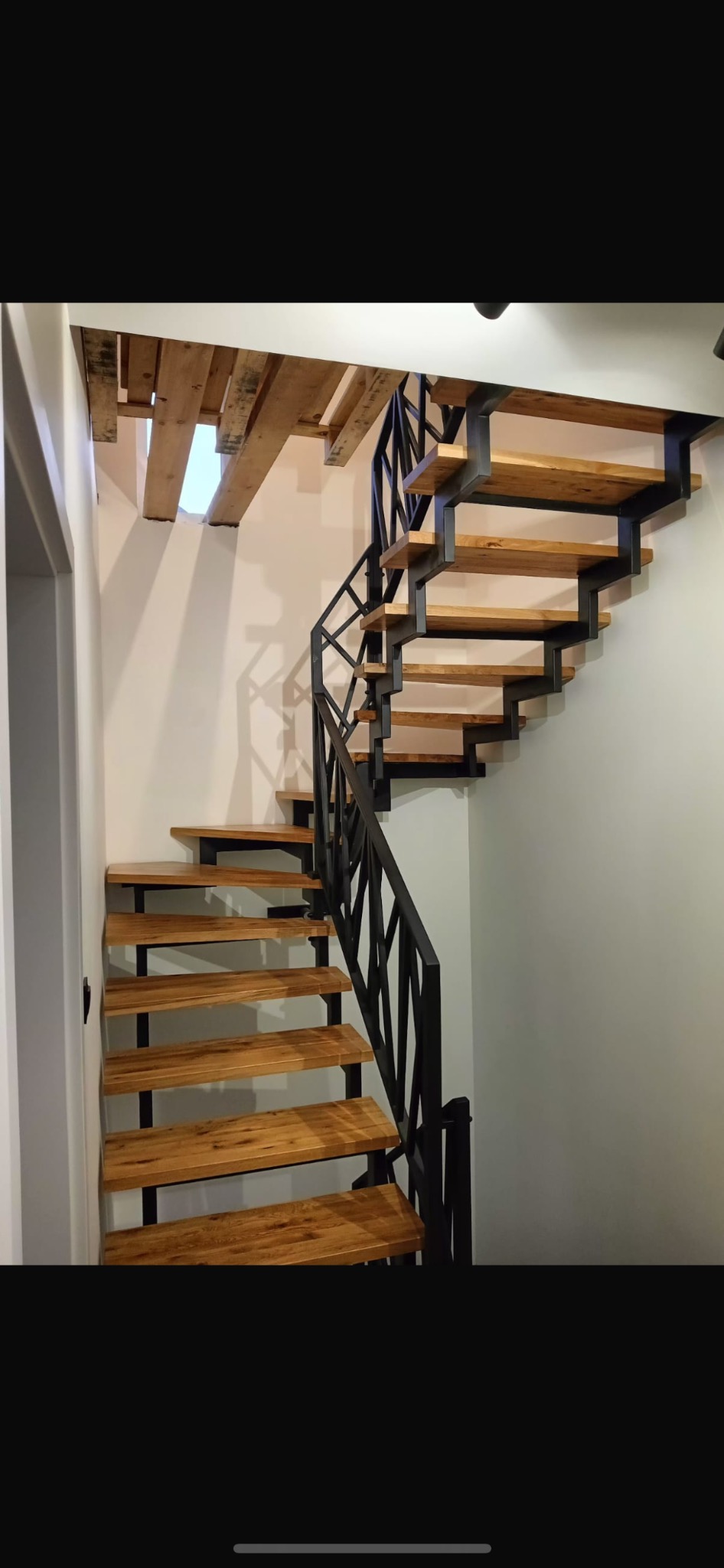 Drewniane schody z czarną, metalową balustradą o geometrycznym wzorze, umieszczone w jasnym wnętrzu. Widoczne drewniane elementy konstrukcyjne pod sufitem.