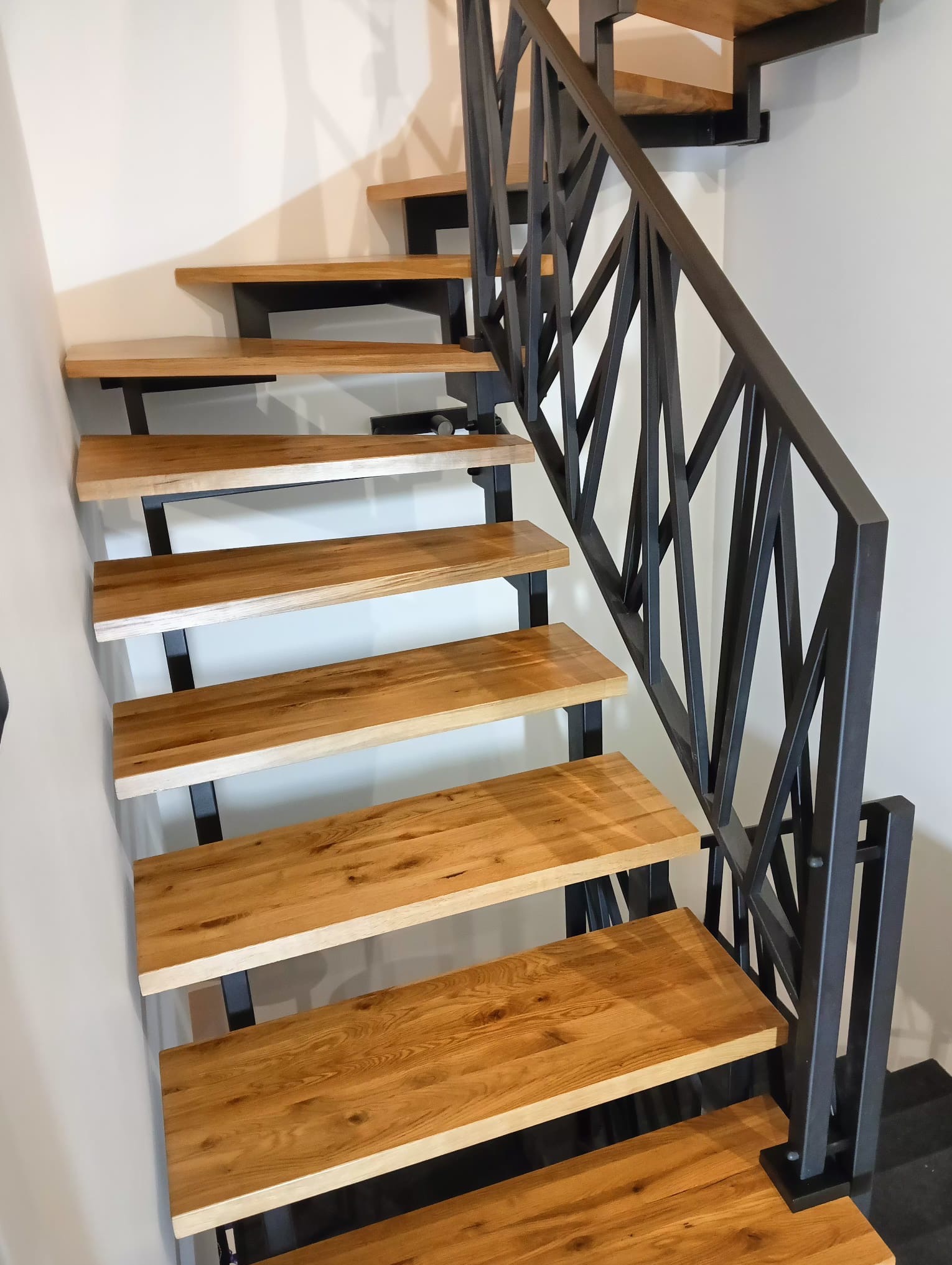 Nowoczesne schody z drewnianymi stopniami i metalową, geometryczną balustradą w kolorze grafitowym, zamontowane przy białej ścianie. Widok z dołu.