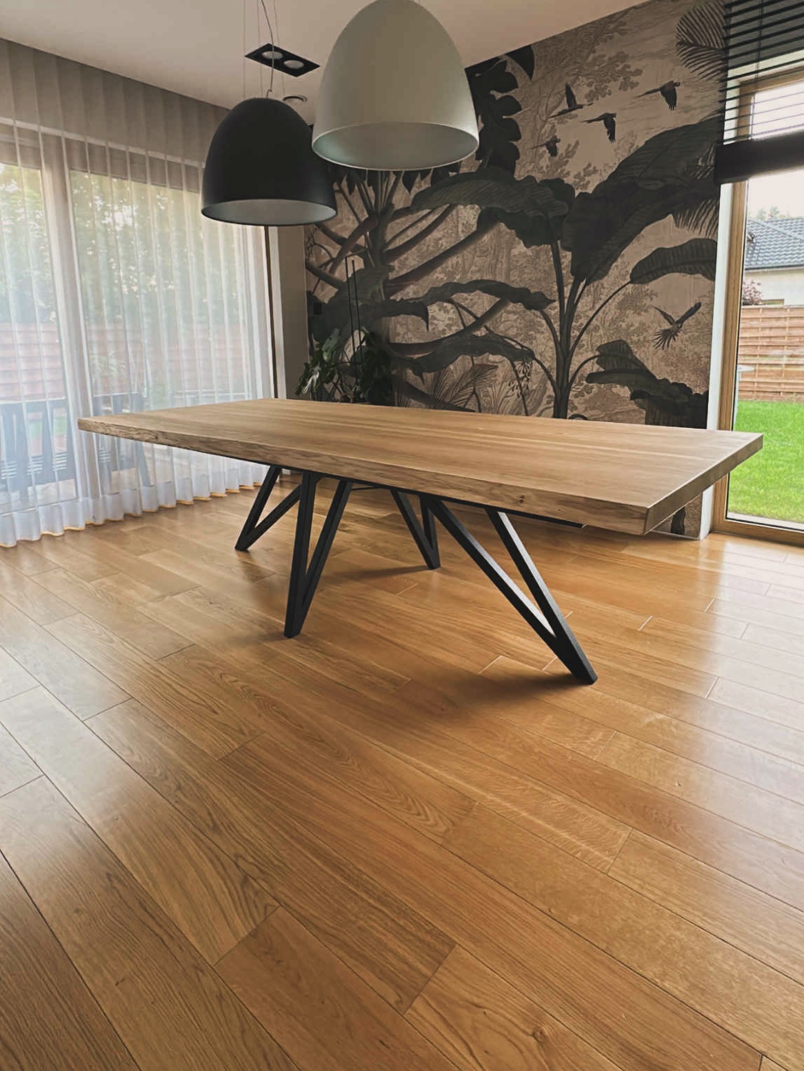 Stół z blatem z jasnego drewna i czarnymi, metalowymi nogami o geometrycznym kształcie. W tle okno z firanką i ściana z motywem roślinnym.