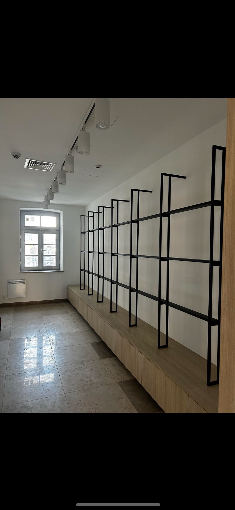 Minimalistyczne, czarne, metalowe regały na jasnym tle ściany, zamontowane nad drewnianą zabudową. Widok na wnętrze z oknem i grzejnikiem.