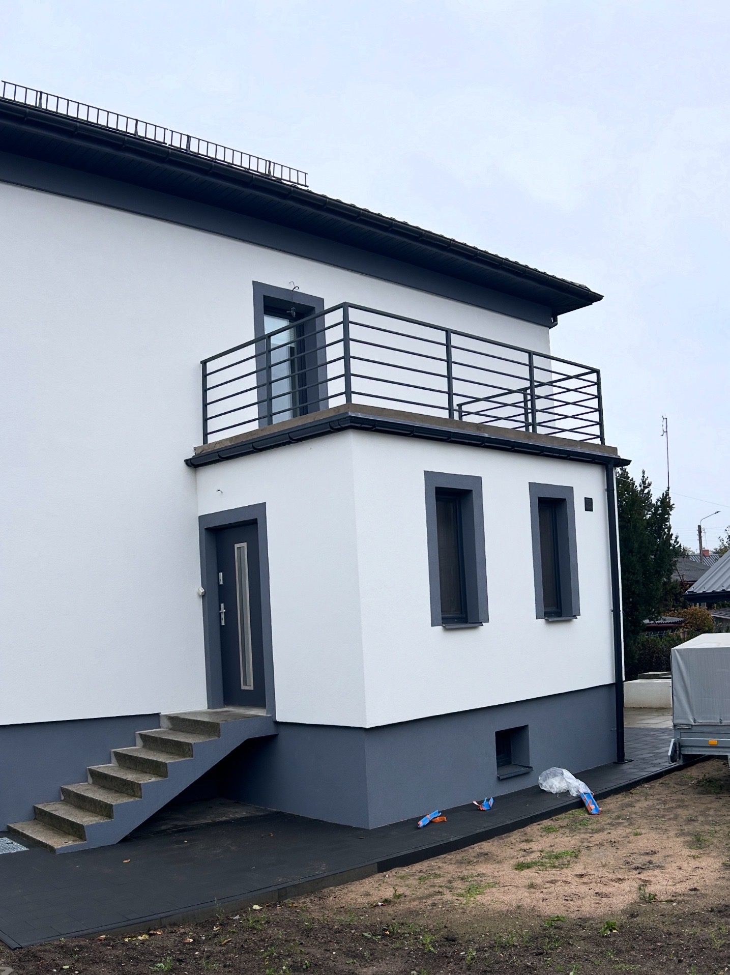 Dwukondygnacyjny dom z balkonem zabezpieczonym czarną, metalową balustradą. Widoczne schody wejściowe i fragment posesji z przyczepą.