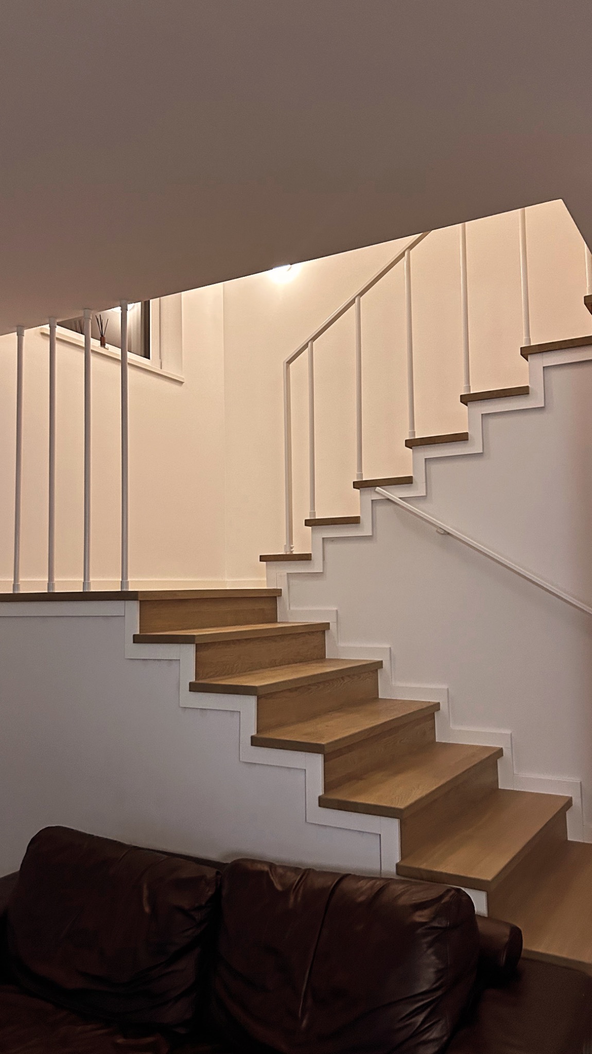Nowoczesne schody drewniane z białymi elementami konstrukcyjnymi i metalową balustradą, widoczne z dołu, z fragmentem sofy w dolnej części kadru.