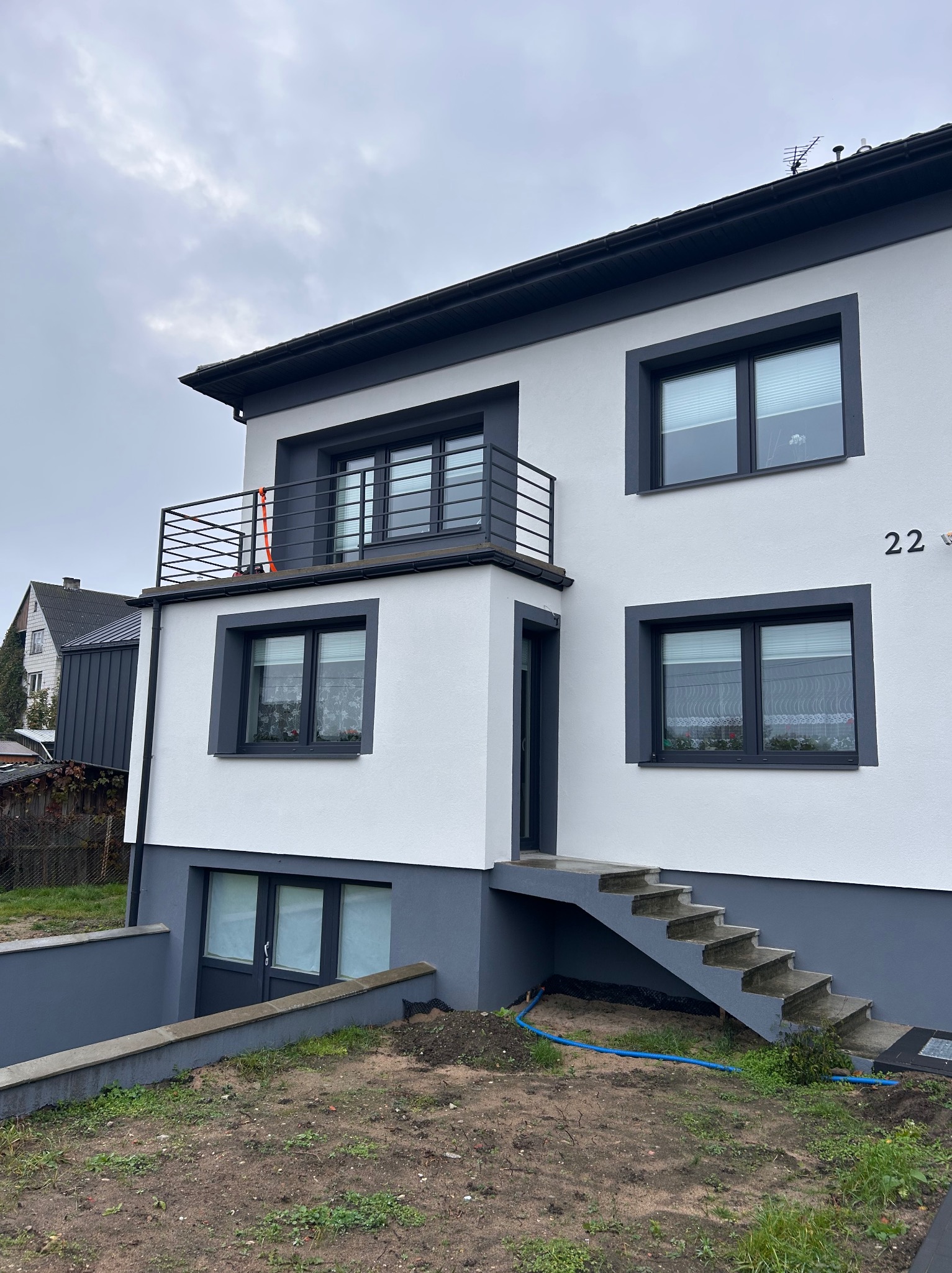 Dwukondygnacyjny dom z balkonem z czarną, metalową balustradą. Szare obramowania okien i numer domu 22. Schody wejściowe z szarego betonu.