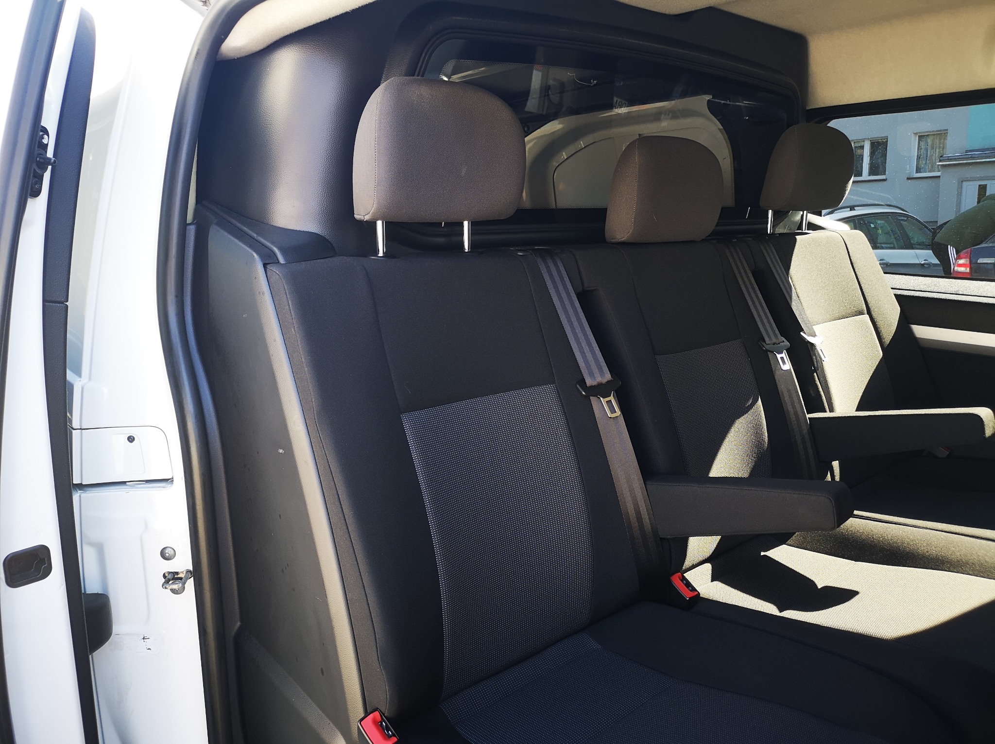 Wnętrze białego busa z trzema fotelami w drugim rzędzie, widoczne pasy bezpieczeństwa i zagłówki. Częściowo widoczne drzwi i budynek w tle.