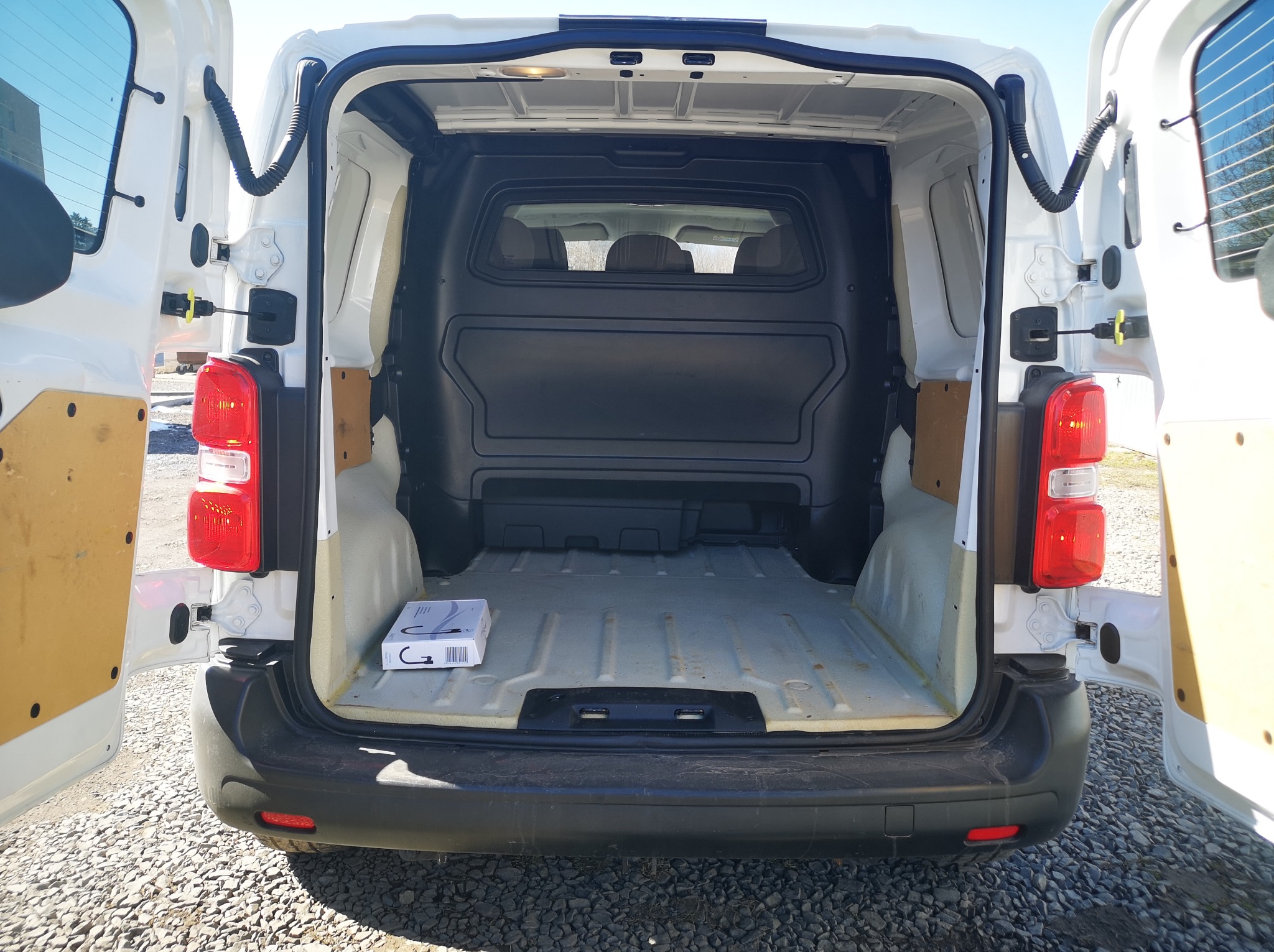 Wnętrze białego busa z otwartymi drzwiami, widok z tyłu. Pusta przestrzeń ładunkowa z pudełkiem w rogu. Widoczne światła i elementy karoserii. Tło: szare kamienie.