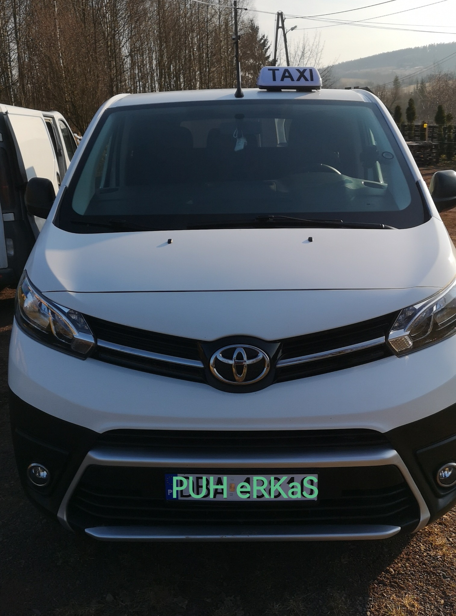 Przód białego busa Toyota z napisem TAXI na dachu, zaparkowanego na zewnątrz w słoneczny dzień. Widoczny fragment innego busa po lewej stronie.