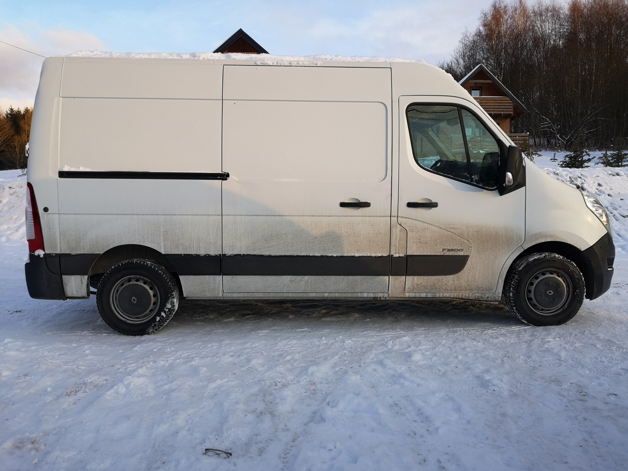 Biały furgon Renault Master zaparkowany na śniegu, widok z boku, zarys drewnianego domu w tle. Lekkie zabrudzenia na karoserii, śnieg na dachu. Zimowy krajobraz.