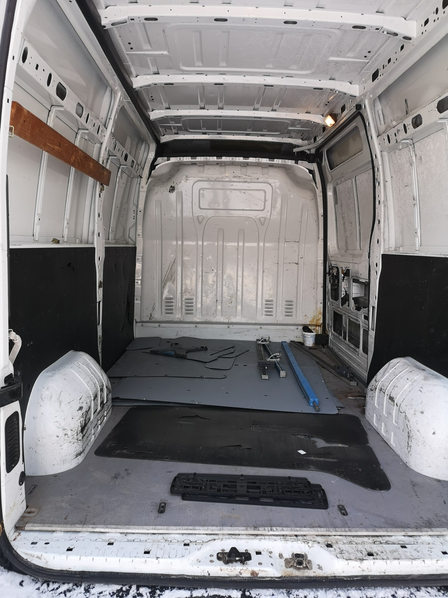 Wnętrze białego busa w trakcie adaptacji: widoczne płyty podłogowe, elementy montażowe i narzędzia. Częściowo wykończone ściany. Prace adaptacyjne w toku.