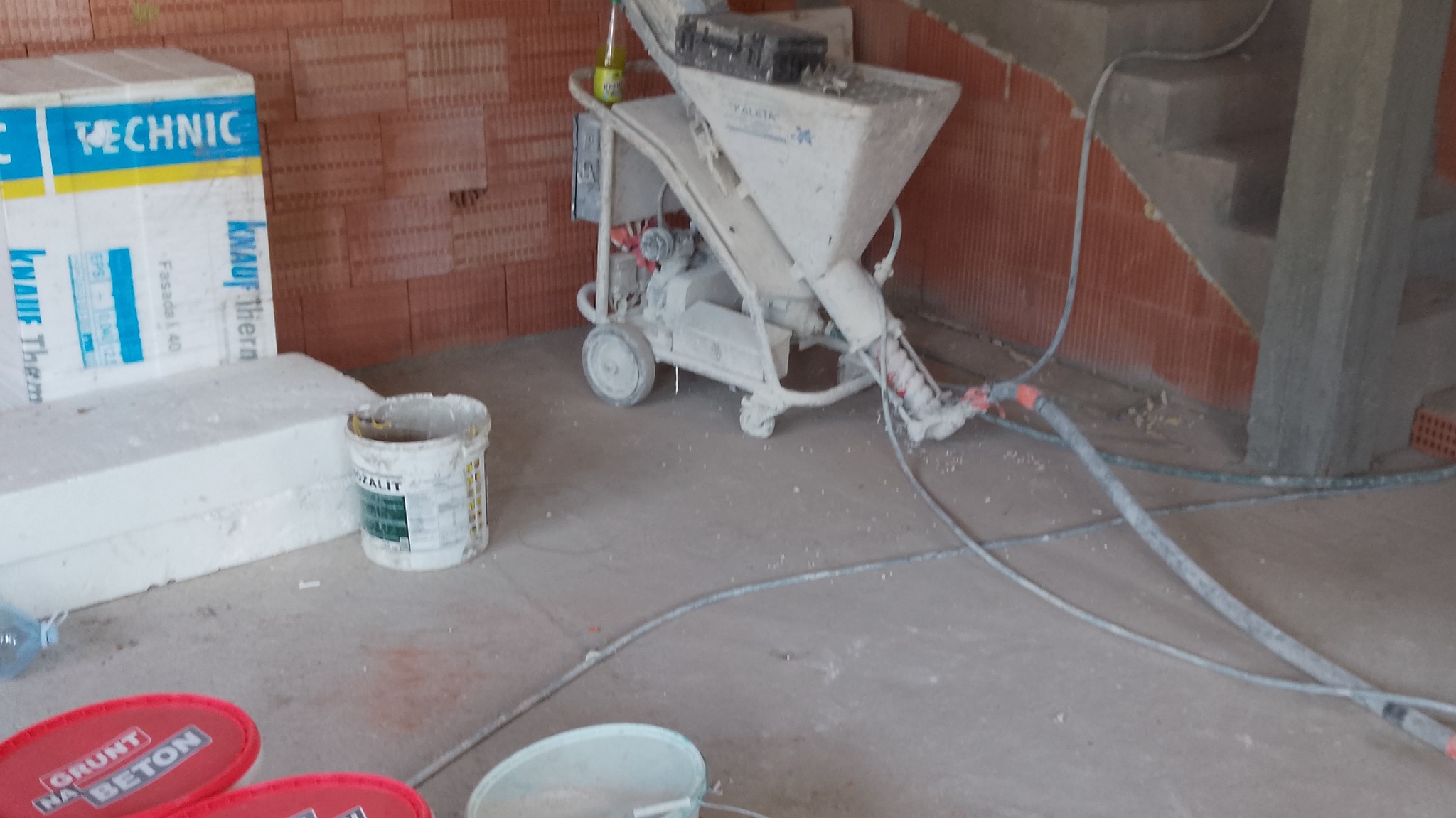 Maszyna do natrysku zaprawy w trakcie pracy, widoczne węże, wiadro z gruntem, styropian. Ceglana ściana w tle. Ujęcie z bliska, z widocznymi detalami.