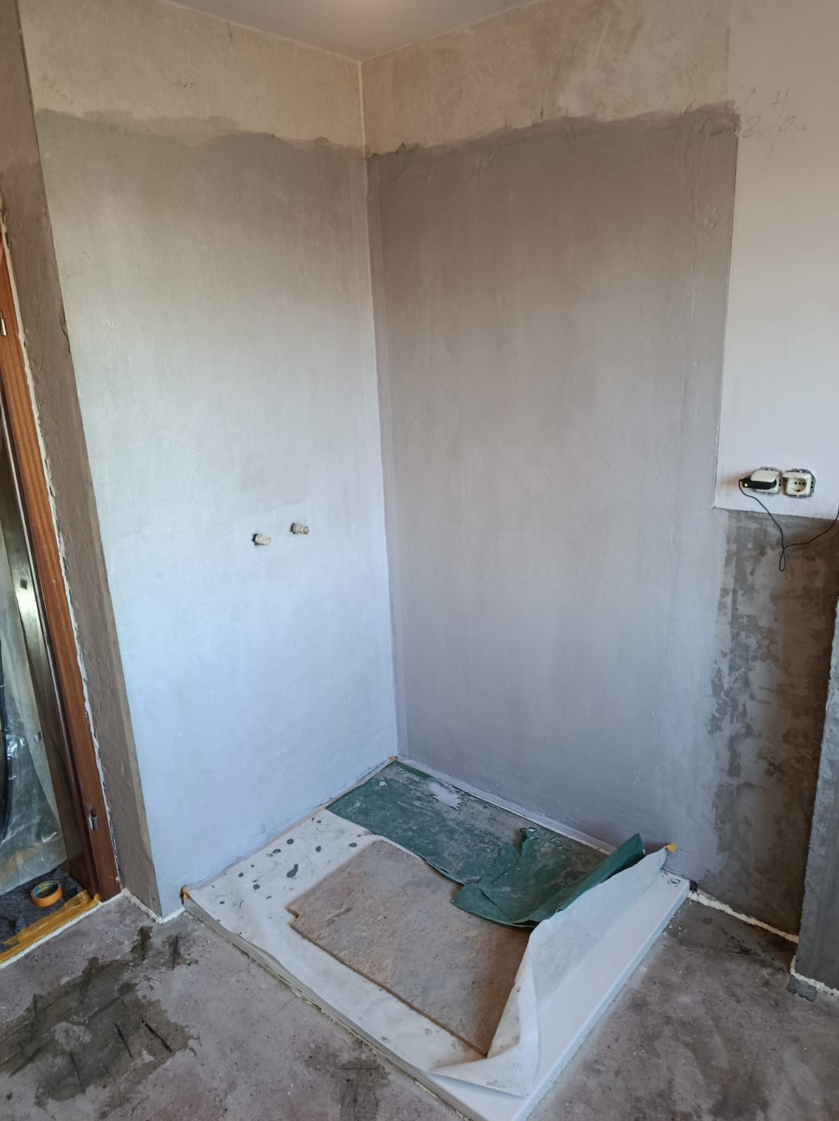 Ściany łazienki po nałożeniu gładzi, przygotowane do dalszych prac wykończeniowych. Widoczne wystające rury i zabezpieczona podłoga. Fragment instalacji elektrycznej.