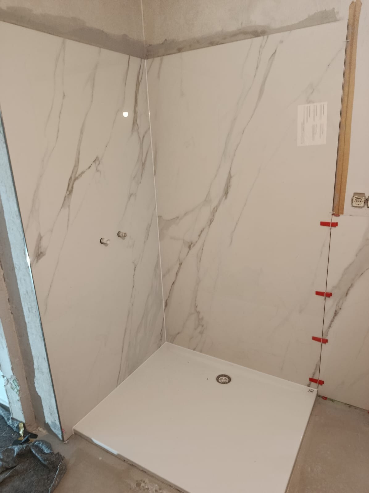 Biała kabina prysznicowa z marmurowymi płytkami w trakcie montażu. Widoczne krzyżyki glazurnicze i niedokończone fugi. Minimalistyczny design.