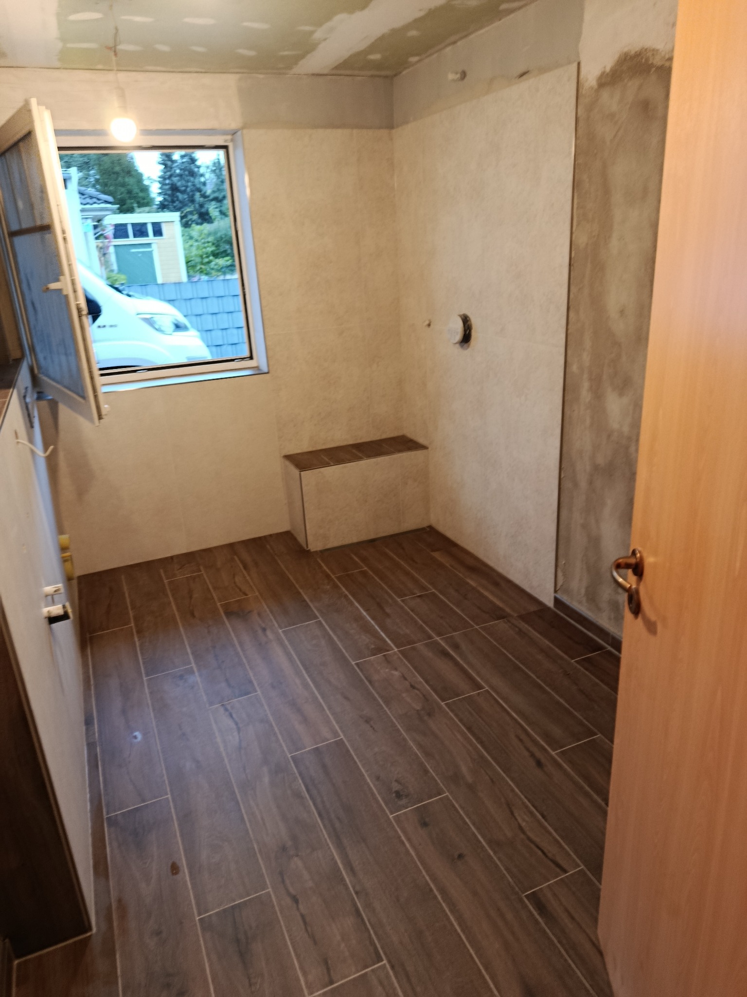 Wnętrze łazienki w trakcie wykańczania: brązowe płytki podłogowe imitujące drewno, jasne płytki ścienne, okno z widokiem na zewnątrz, fragment ściany przygotowany pod płytki.