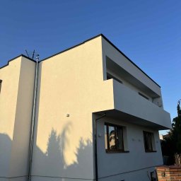 MIT BUD-INSTAL - Nowoczesny dom z jasną elewacją i balkonem, widziany pod kątem. Widoczna metalowa rura spustowa i cień drzewa na ścianie. Błękitne niebo w tle.