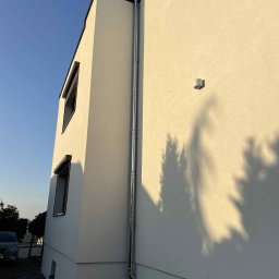 MIT BUD-INSTAL - Elewacja budynku z kominem w Świerklanach, oświetlona popołudniowym słońcem. Widoczne okna z roletami i cień drzewa na ścianie. Nowoczesny styl.