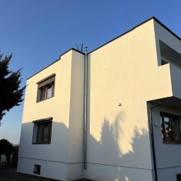 MIT BUD-INSTAL - Nowoczesny, dwukondygnacyjny dom z białą elewacją i brązowymi oknami. Widoczna metalowa rura pionowa na elewacji. Czyste, błękitne niebo w tle. Cień drzewa na ścianie.