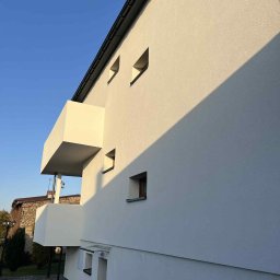 MIT BUD-INSTAL - Biała elewacja budynku z balkonem w kształcie prostopadłościanu, oświetlona popołudniowym słońcem, z małymi, kwadratowymi oknami i czarną rynną. Widok z dołu.