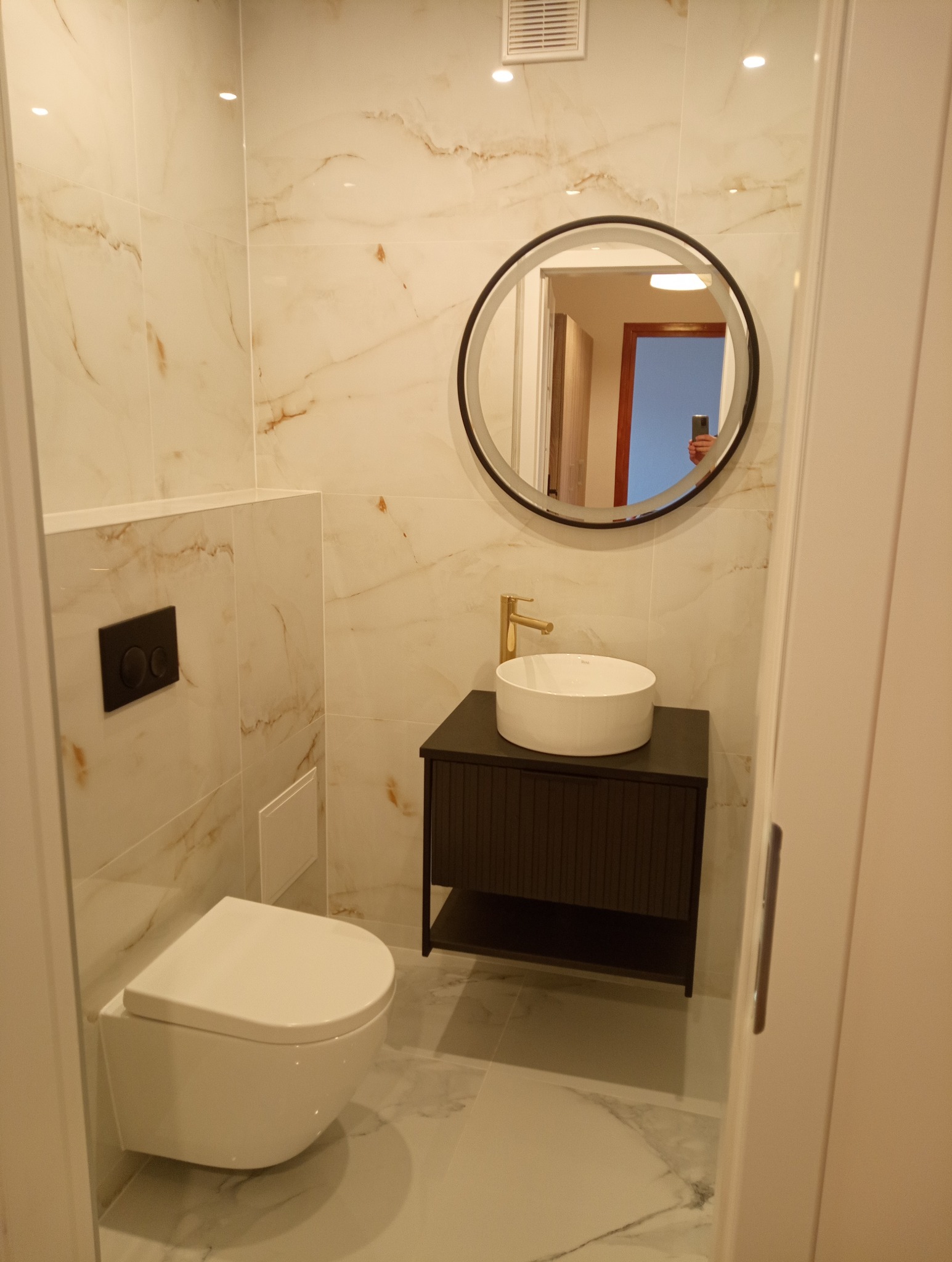 Elegancka łazienka z marmurowymi płytkami, okrągłym lustrem z podświetleniem, wiszącą toaletą i umywalką na szafce z złotą armaturą. Widok na wnętrze przez odbicie w lustrze.