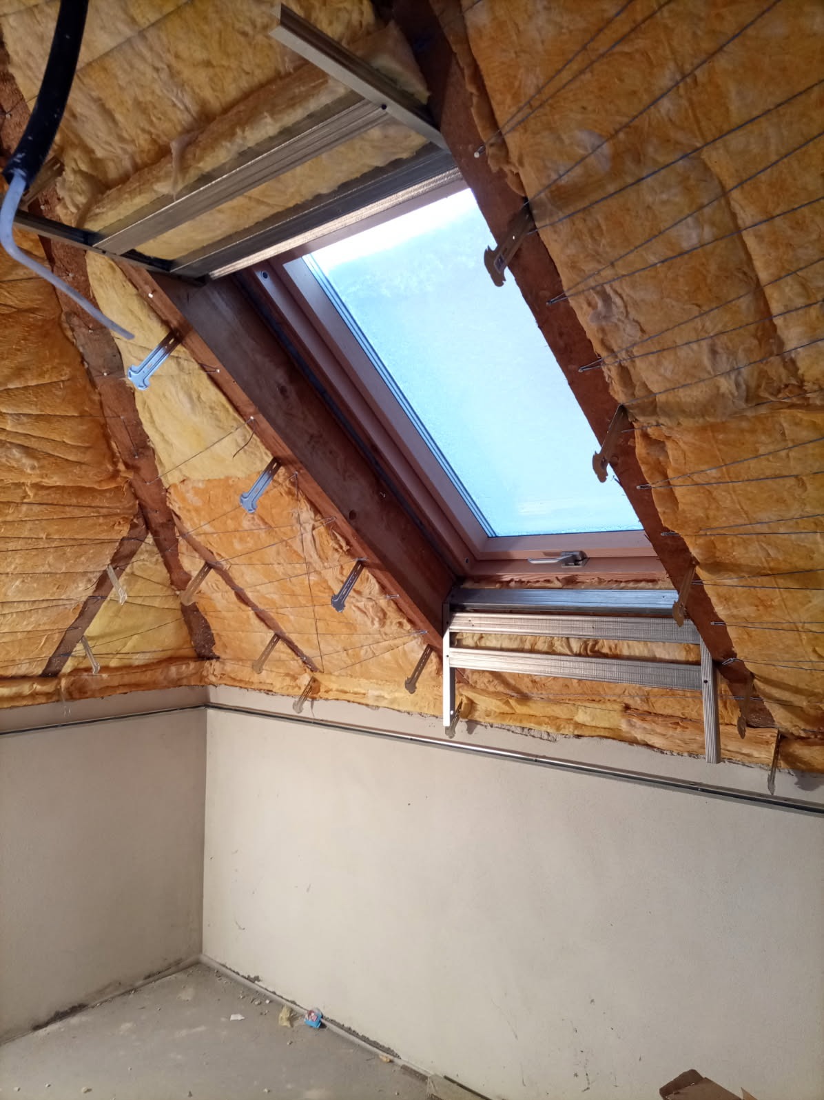 Poddasze z oknem dachowym w trakcie izolacji. Widoczne metalowe profile i wełna mineralna. Ściany przygotowane do dalszych prac wykończeniowych.