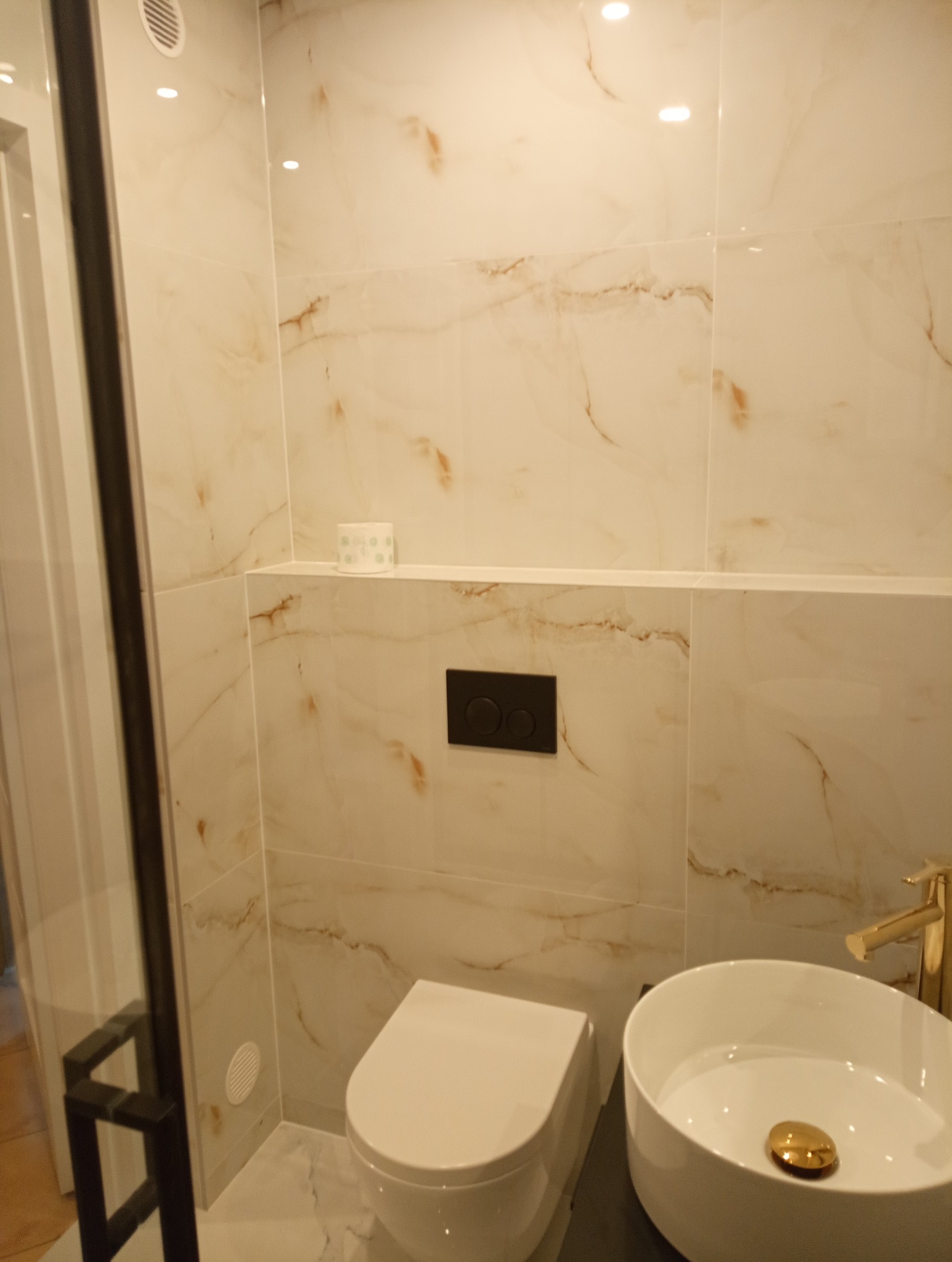 Wykończona łazienka z beżowymi płytkami imitującymi marmur, podwieszana toaleta i umywalka nablatowa z baterią w kolorze złotym. Widoczna kabina prysznicowa.