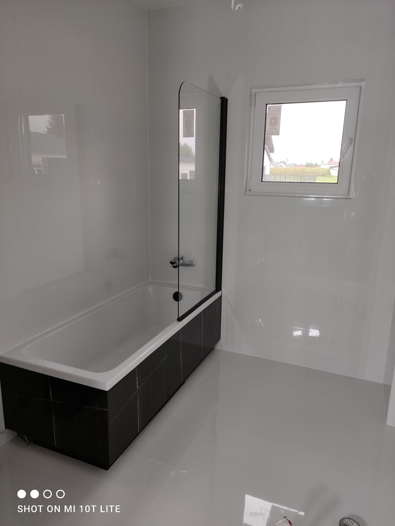 Nowoczesna łazienka z wanną obudowaną czarnymi płytkami z białymi żyłami, szklanym parawanem i jasnymi ścianami wykończonymi panelami.