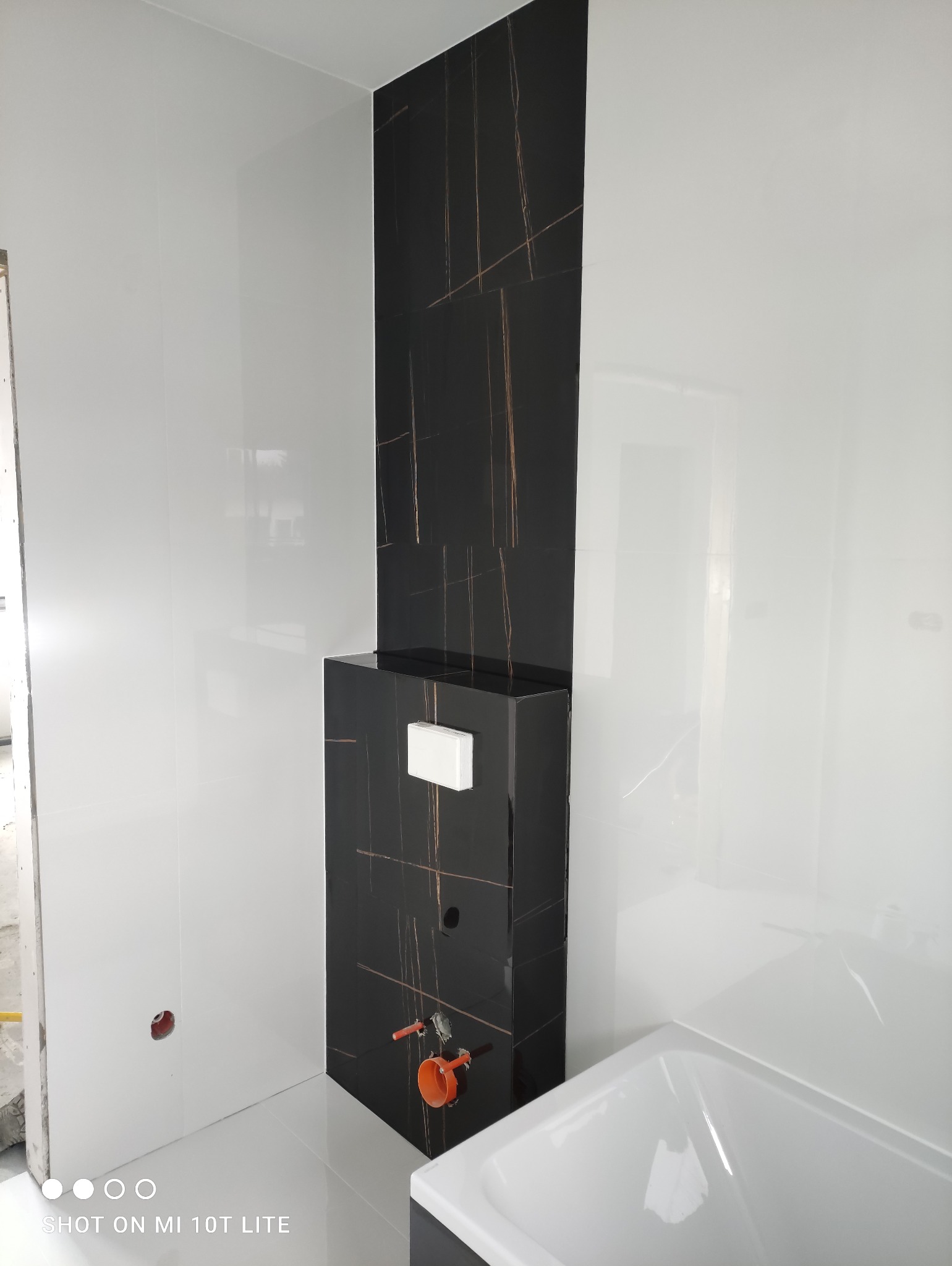 Błyszczące, białe i czarne płytki w łazience, z widoczną instalacją podtynkową WC i fragmentem wanny. Minimalistyczny design, nowoczesne wykończenie.