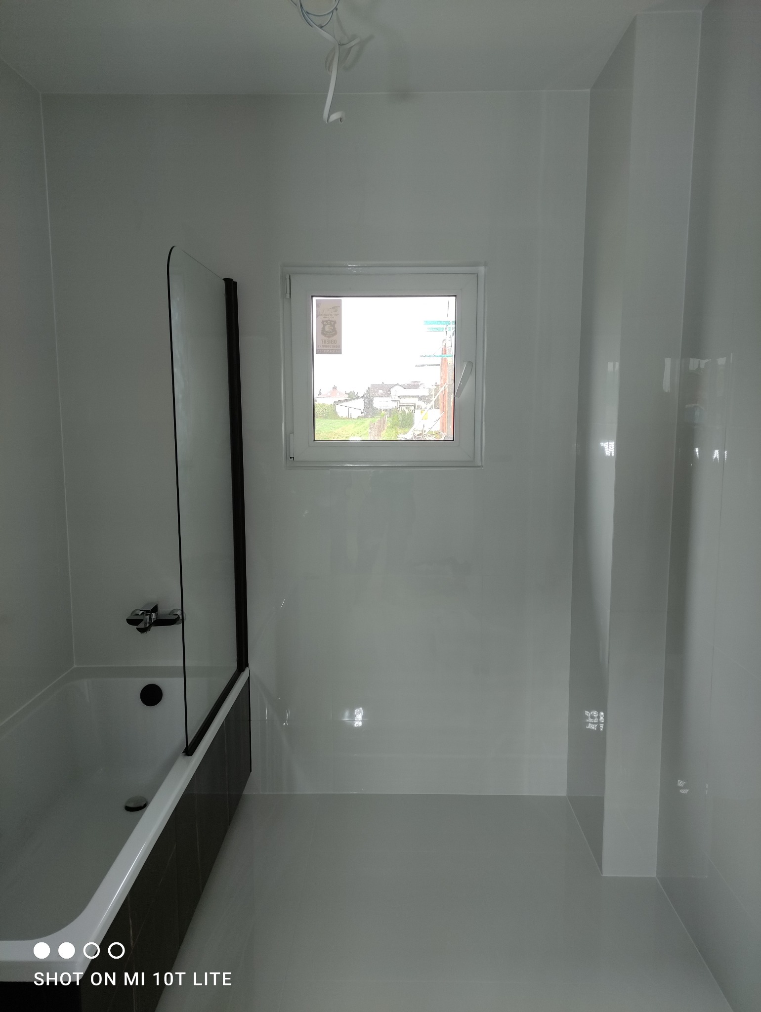 Biała łazienka po remoncie z wanną z parawanem, małym oknem i połyskującymi płytkami na ścianach i podłodze. Widoczne wystające kable.