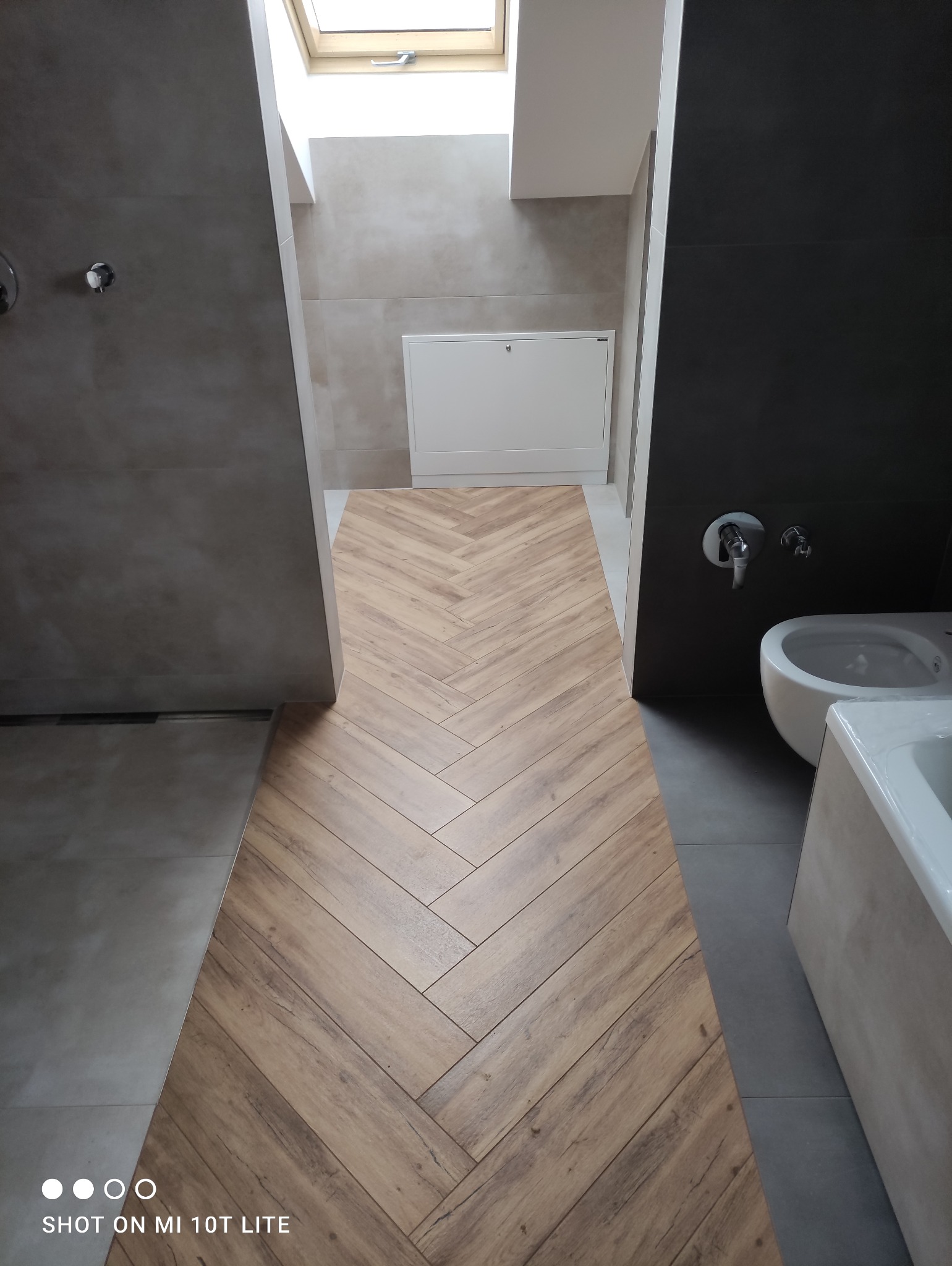 Wykończona łazienka z drewnopodobną podłogą ułożoną w jodełkę, szarą glazurą i oknem dachowym. Widoczna wanna, bidet i szafka rewizyjna.