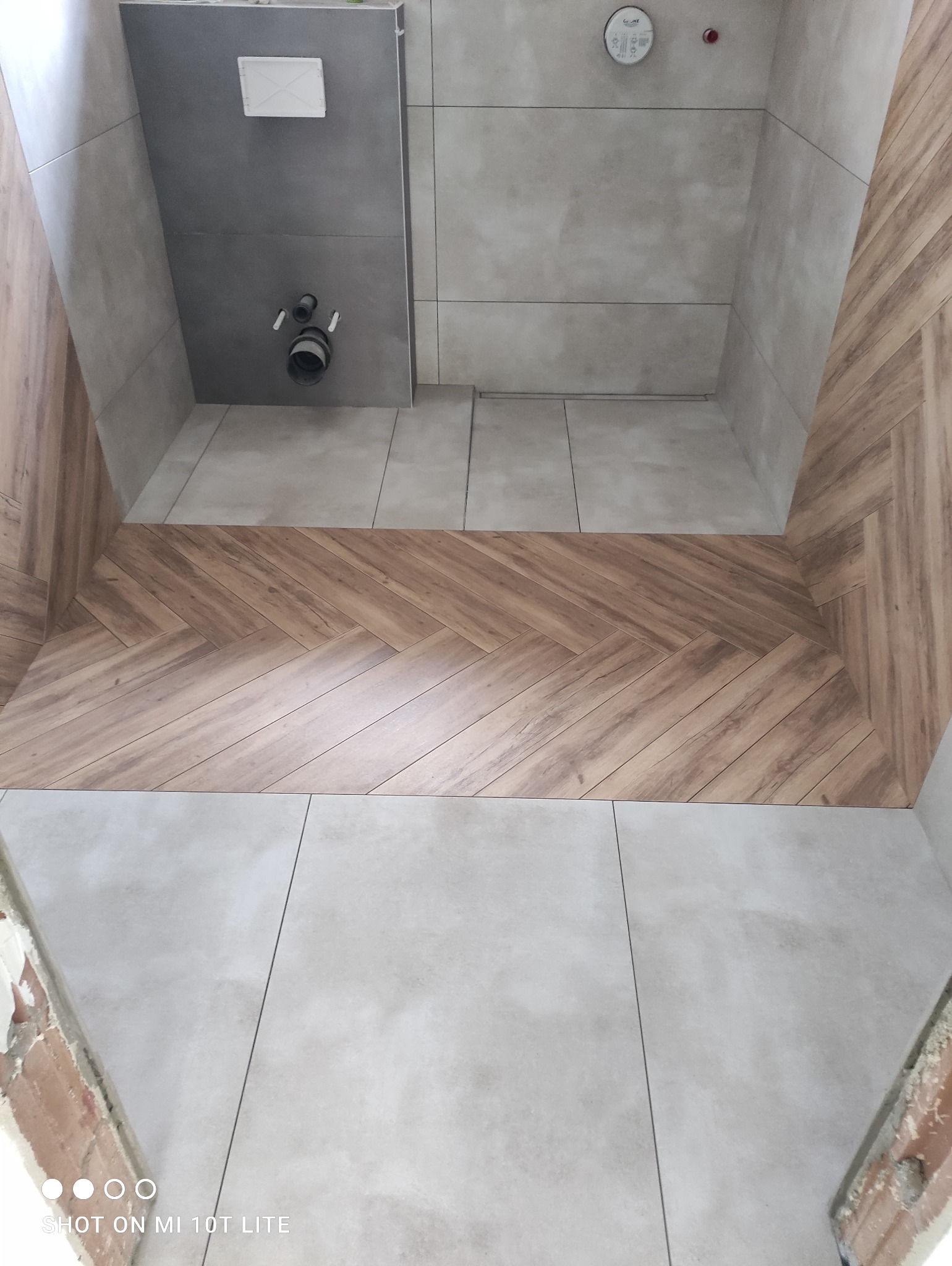 Wykończona łazienka z drewnopodobną glazurą ułożoną w jodełkę na ścianie, jasne płytki podłogowe i zabudowa WC z szarą glazurą. Widok z góry.