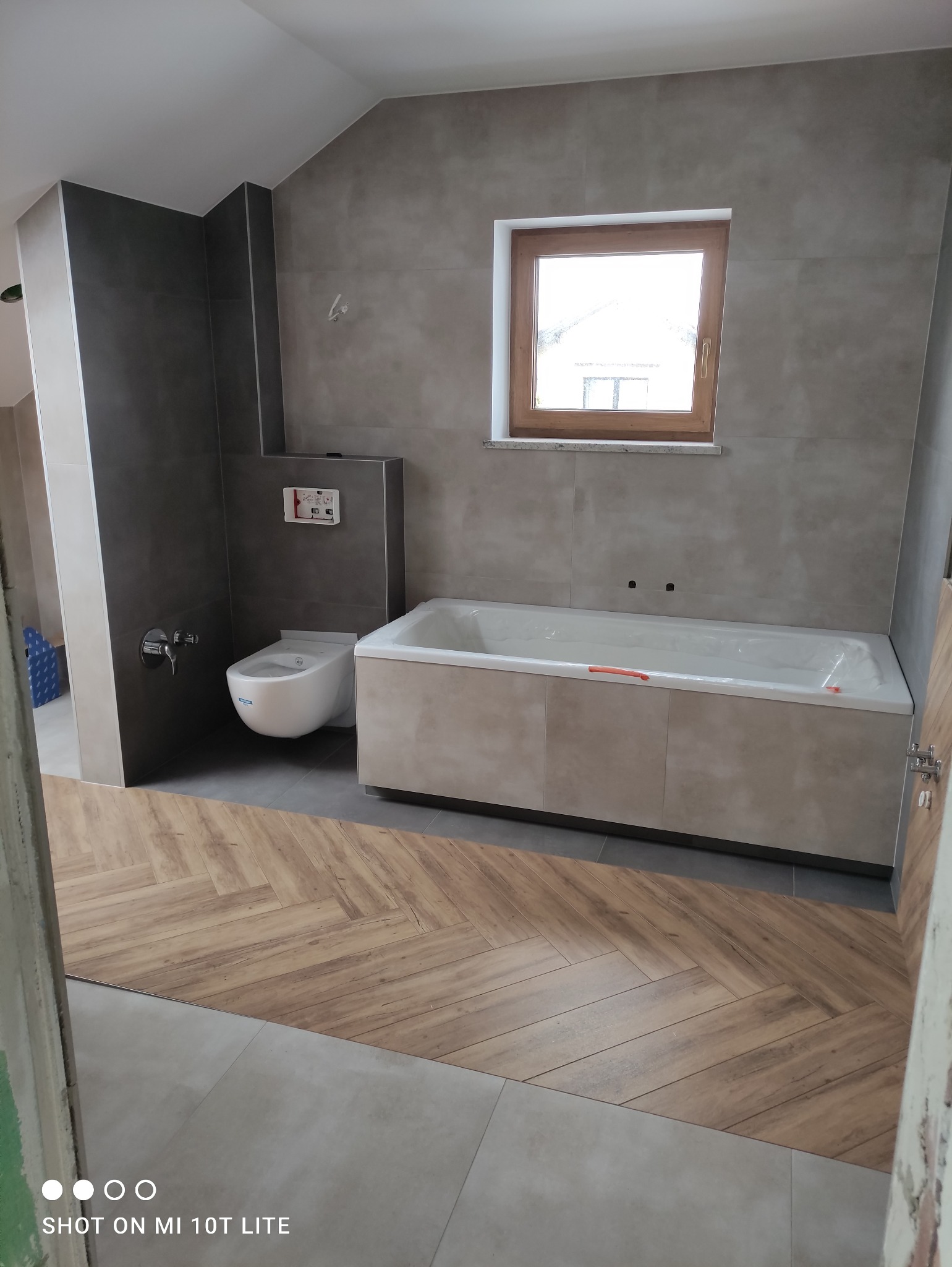 Nowoczesna łazienka z szarymi płytkami, drewnopodobną podłogą ułożoną w jodełkę i oknem z drewnianą ramą. Uwagę zwraca minimalistyczna toaleta i zabudowana wanna.