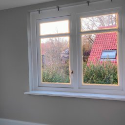 Kraw-Bud - Świeżo pomalowane wnętrze z oknem na zewnątrz, ściany w odcieniach szarości, biała rama okienna i parapet. Widok na dom z czerwoną dachówką.