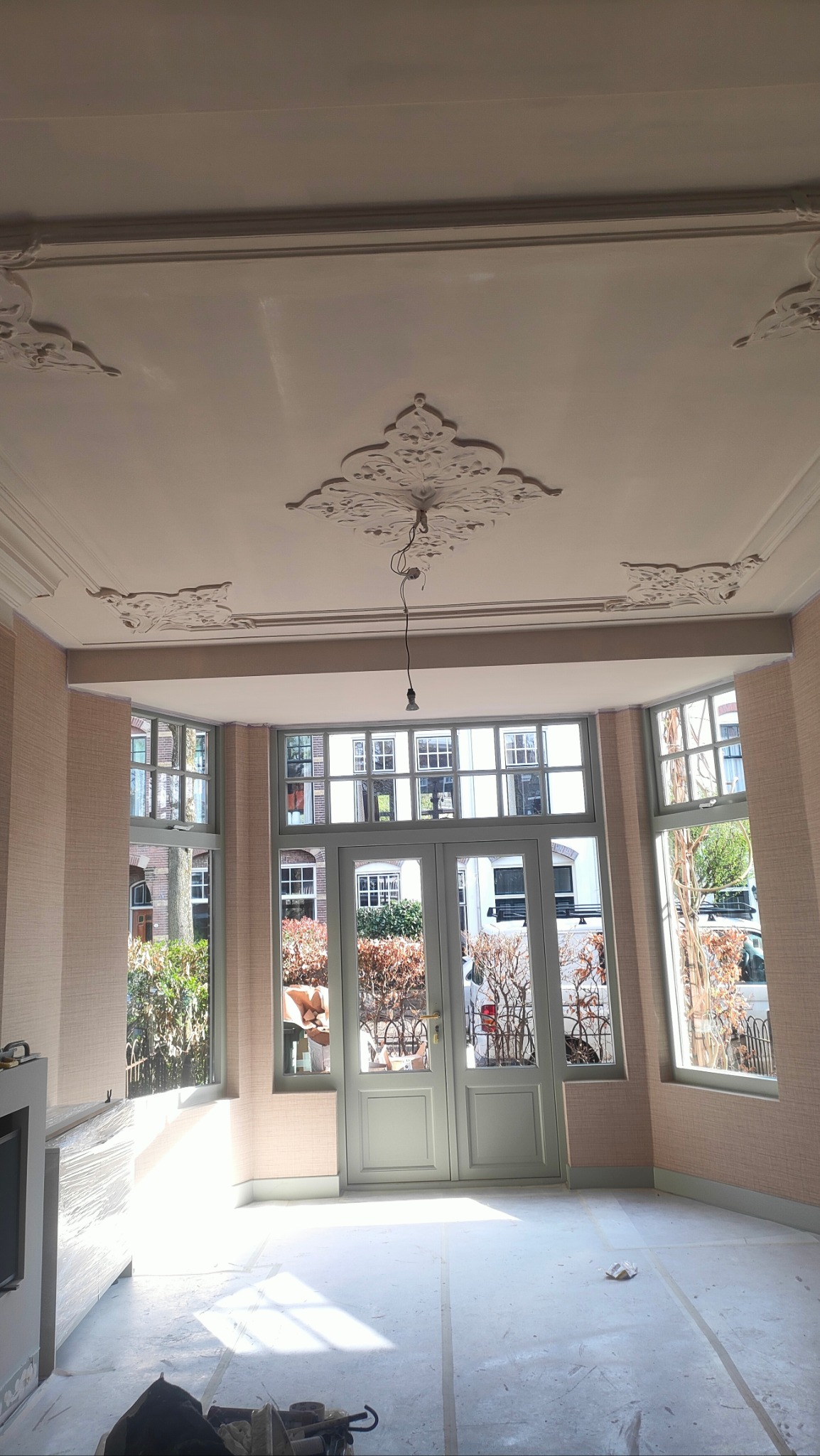 Jasne wnętrze z dekoracyjnym sufitem i dużymi oknami. Ściany w delikatnym odcieniu różu, podłoga przygotowana do dalszych prac wykończeniowych. Widok na zieleń za oknem.