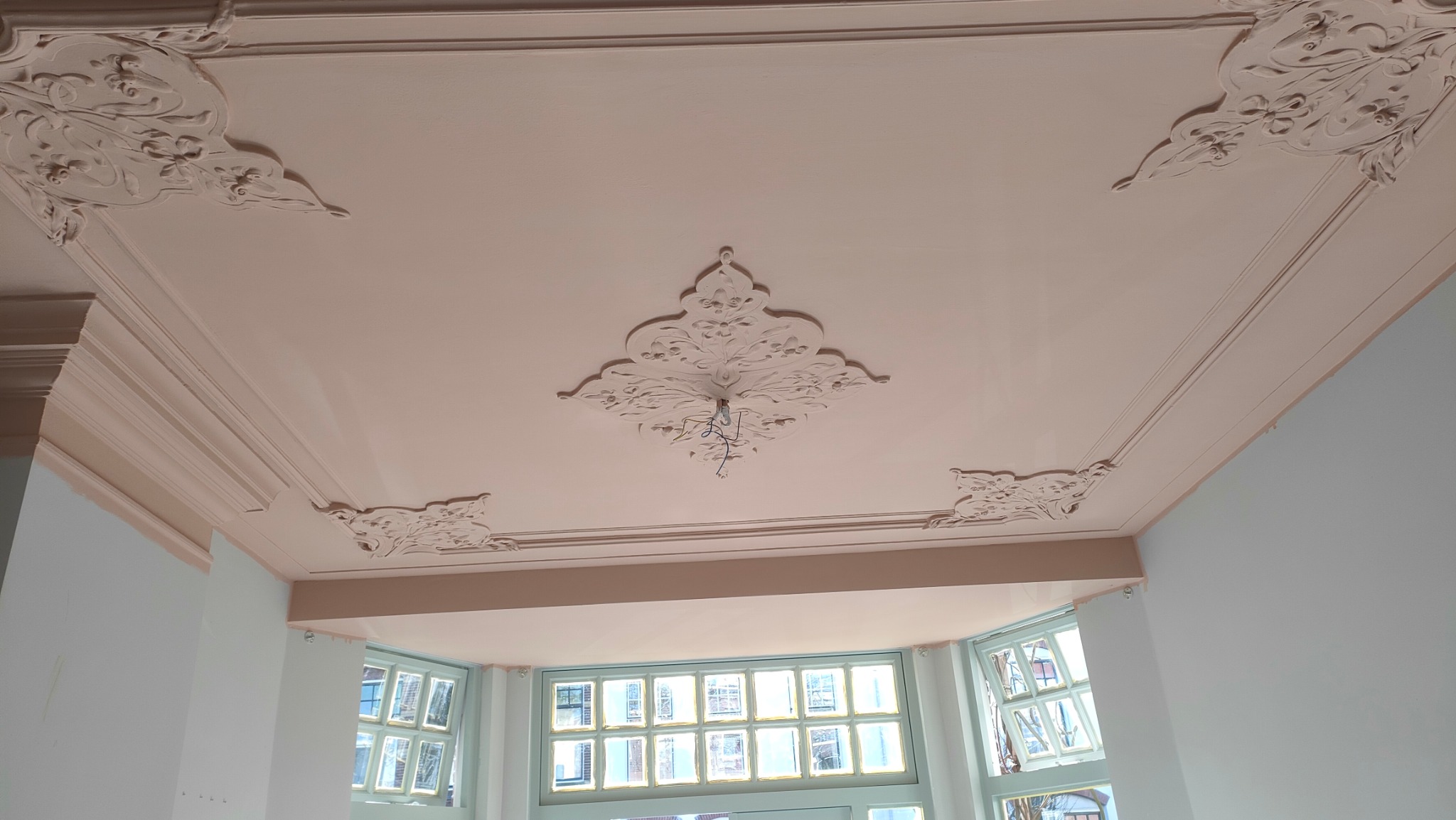 Jasnoróżowy sufit z dekoracyjnymi sztukateriami i listwami, świeżo po malowaniu, widok z dołu na okno z szybami w kratkę.