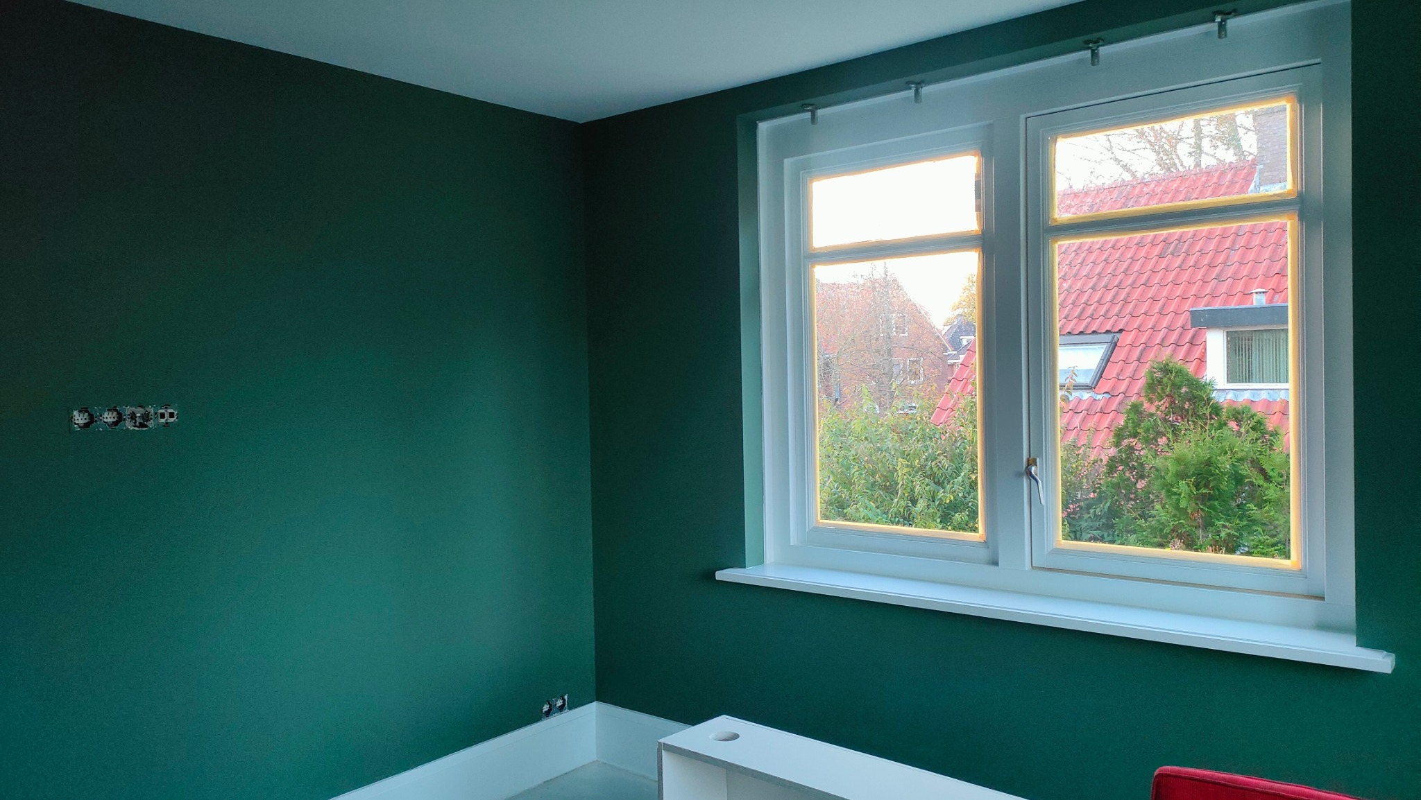 Pokój po malowaniu na butelkową zieleń z białym oknem, widok na domy i zieleń za oknem. Gniazdka elektryczne widoczne na ścianie.
