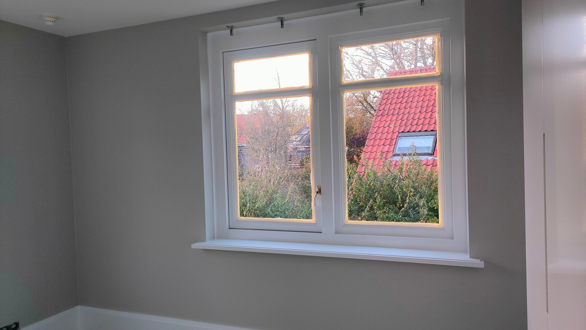Świeżo pomalowane wnętrze z oknem na zewnątrz, ściany w odcieniach szarości, biała rama okienna i parapet. Widok na dom z czerwoną dachówką.