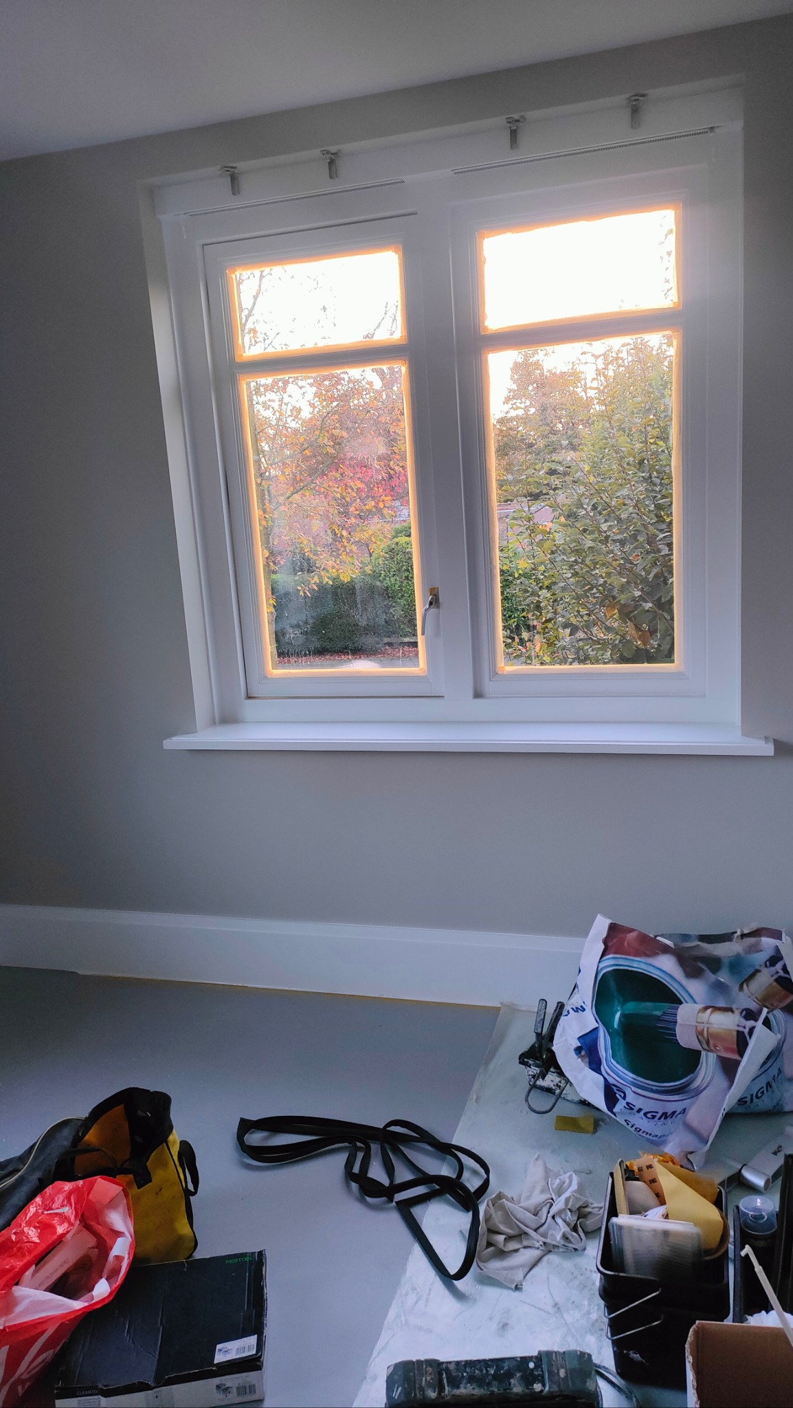 Poddasze po malowaniu: białe okno z widokiem na jesienny ogród, ściany w odcieniach szarości, narzędzia malarskie i torba robocza na podłodze.