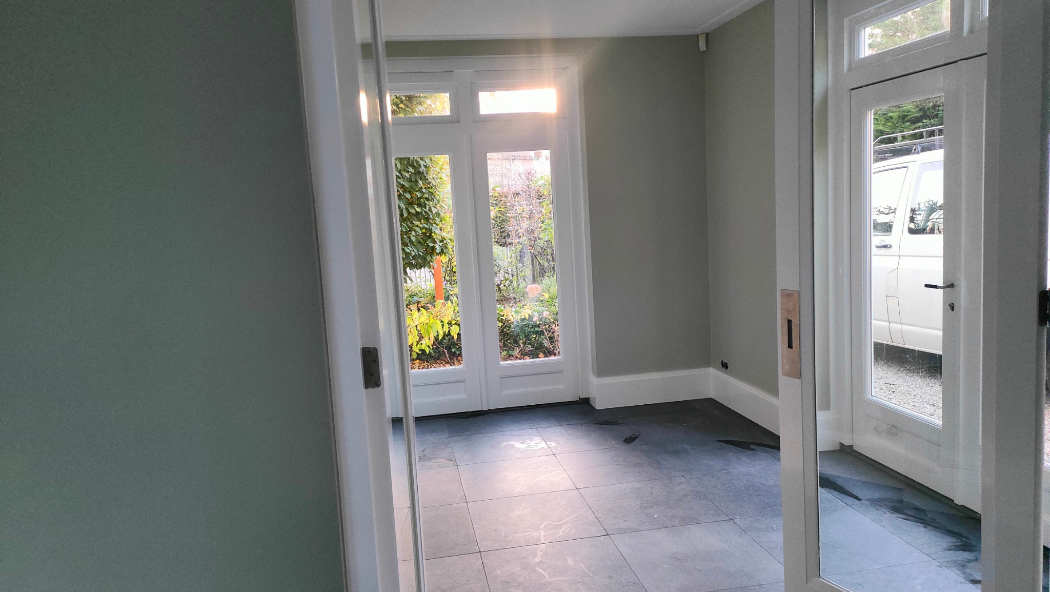 Pomalowane na szaro wnętrze z widokiem na ogród przez białe, przeszklone drzwi. Podłoga wyłożona ciemnymi płytkami. Widoczny fragment białego samochodu.