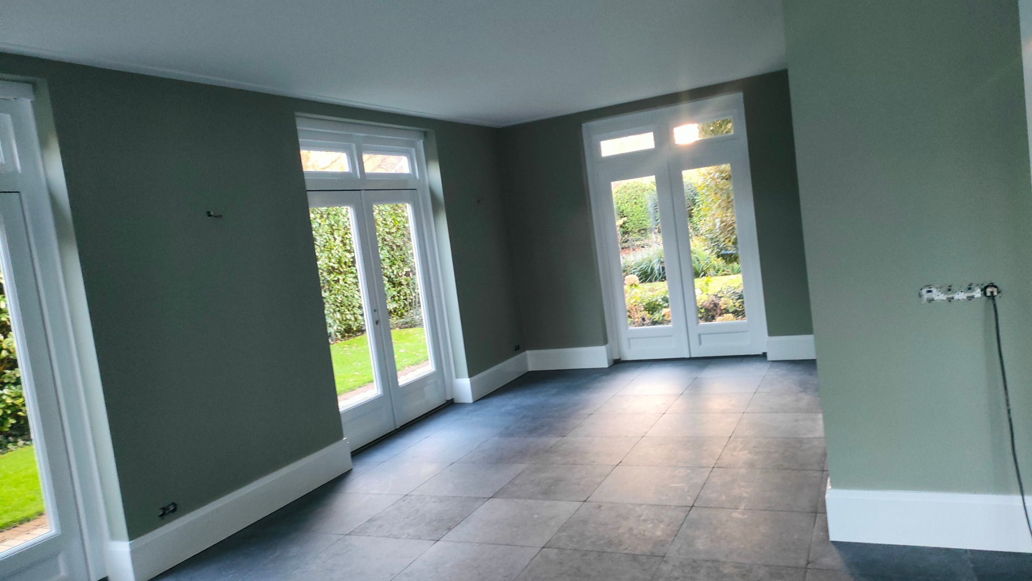 Pomieszczenie po malowaniu ścian na oliwkowy kolor, z widocznymi białymi, przeszklonymi drzwiami balkonowymi i szarą podłogą z płytek. Widoczne gniazdka elektryczne.