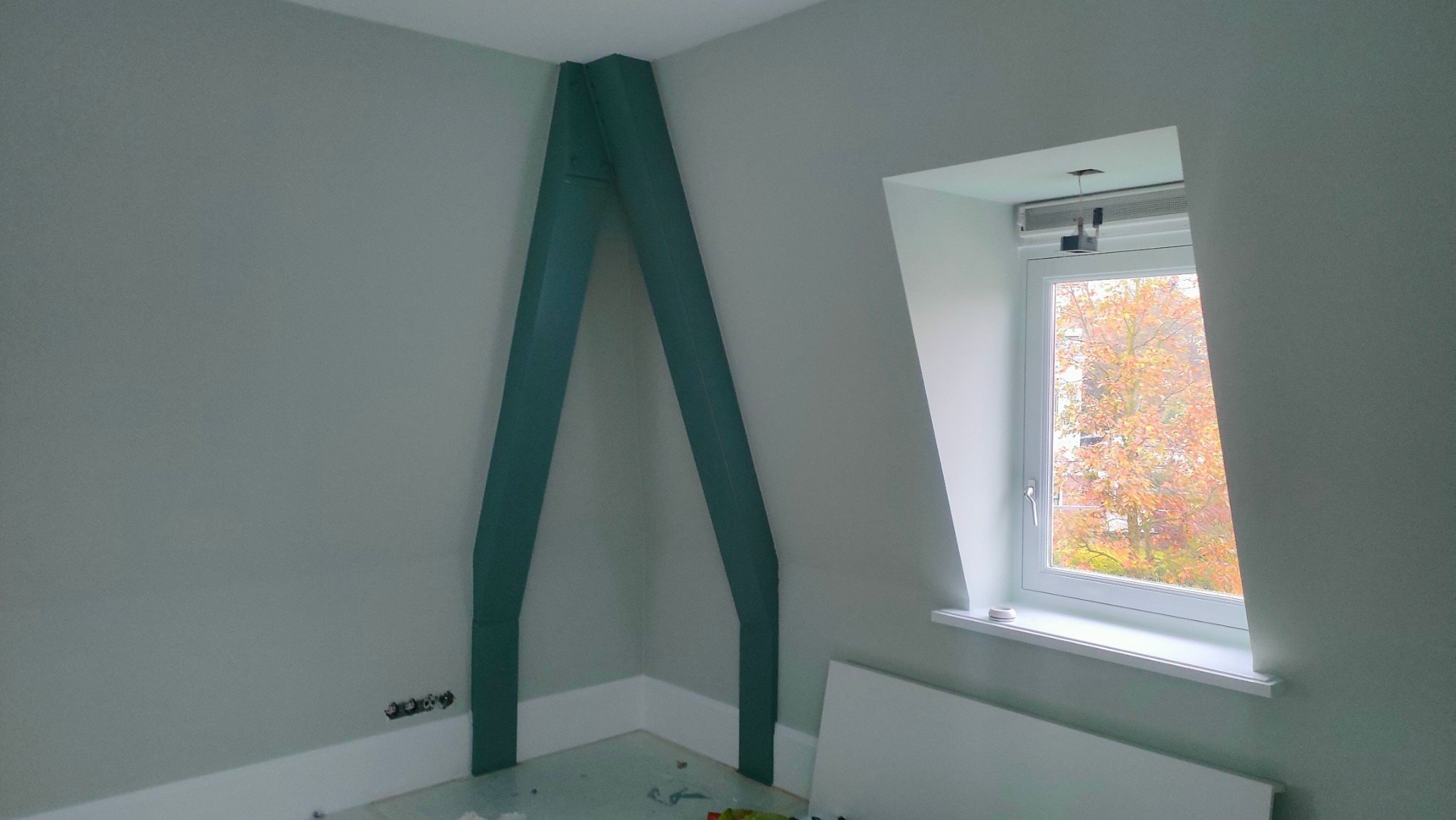 Pomalowane na zielono wzmocnienie konstrukcyjne poddasza w rogu pokoju, ściany w odcieniach szarości, białe okno z widokiem na jesienne drzewo.