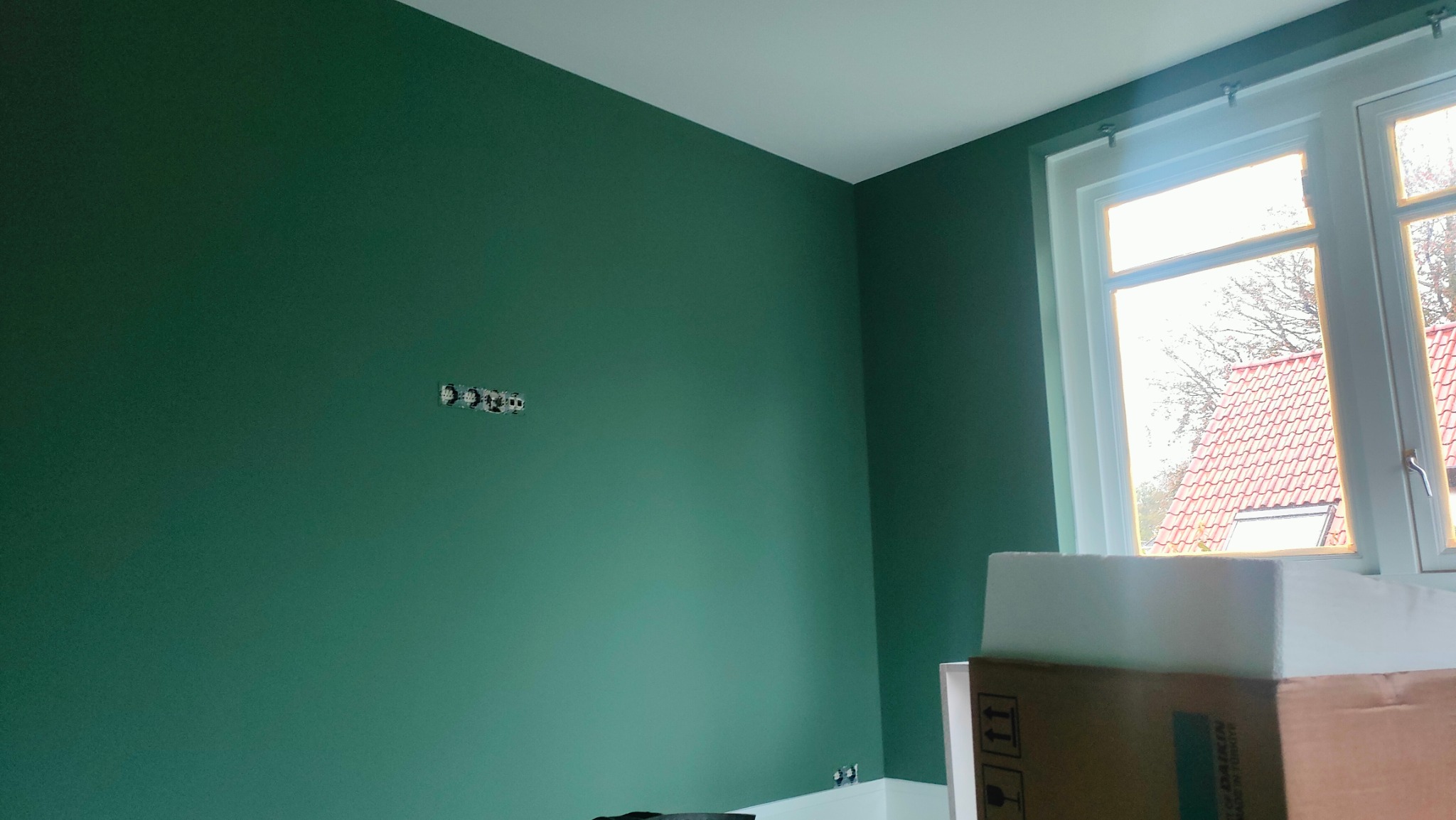 Ściana pomalowana na zielono z widocznymi puszkami elektrycznymi i oknem w białej ramie. Częściowo widoczne kartony z materiałami budowlanymi.