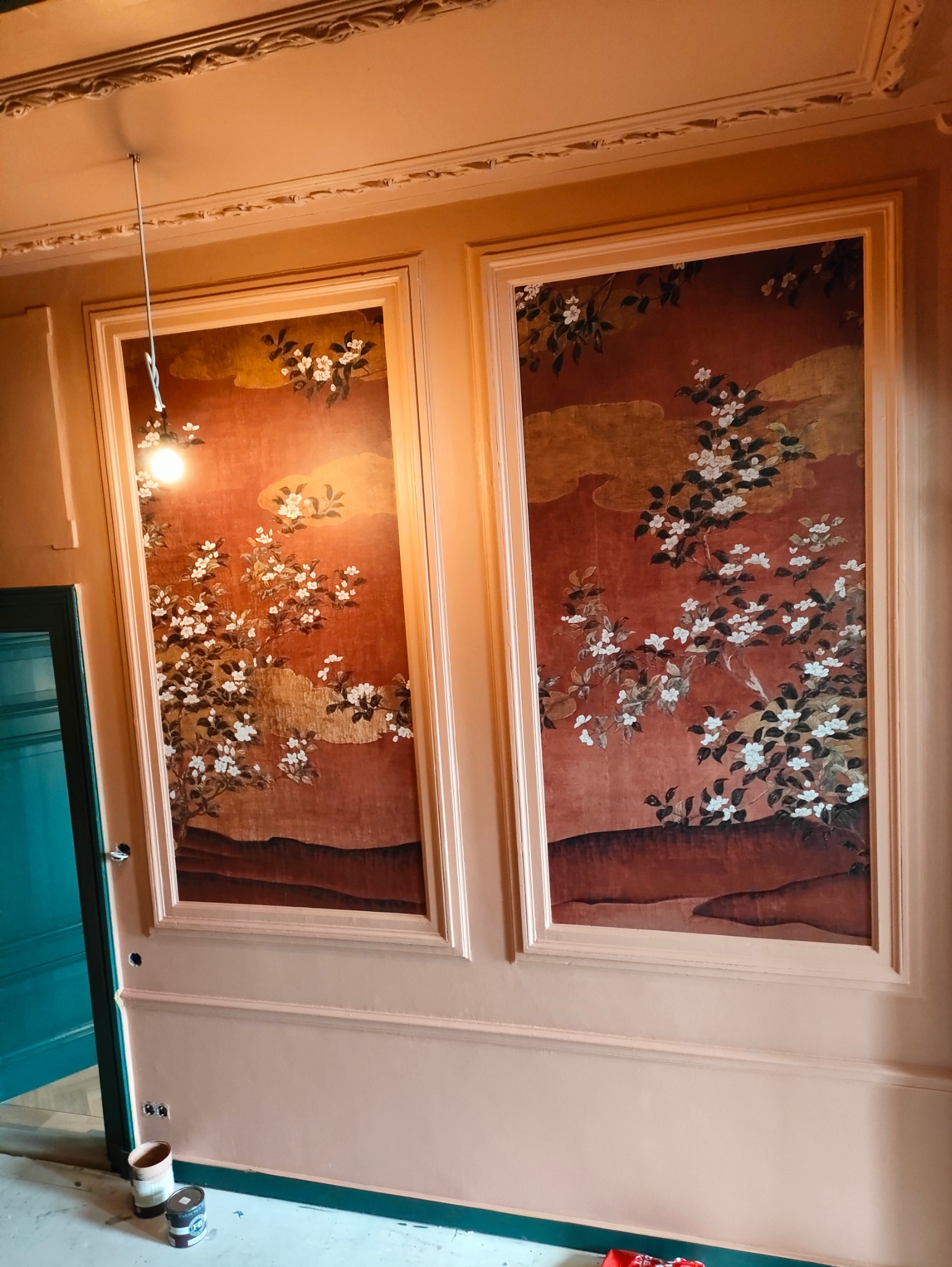 Wykończona ściana z dwoma dekoracyjnymi panelami w stylu orientalnym, z motywem kwitnących gałęzi na tle w odcieniach czerwieni i złota. Widoczne puszki z farbą.