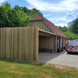 BEBEK, BEBEK MAN BUŁKA I SPÓŁKA DARIUSZ LENART - Drewniana konstrukcja w ogrodzie z pionowymi panelami i częściowo zadaszonym miejscem parkingowym, widoczny pracownik na dachu, samochód dostawczy w tle.