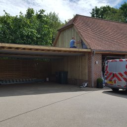 BEBEK, BEBEK MAN BUŁKA I SPÓŁKA DARIUSZ LENART - Konstrukcja zadaszenia garażowego w trakcie budowy, widoczny pracownik na dachu, drewniane elementy i ceglana ściana budynku w tle, zaparkowany samochód dostawczy.